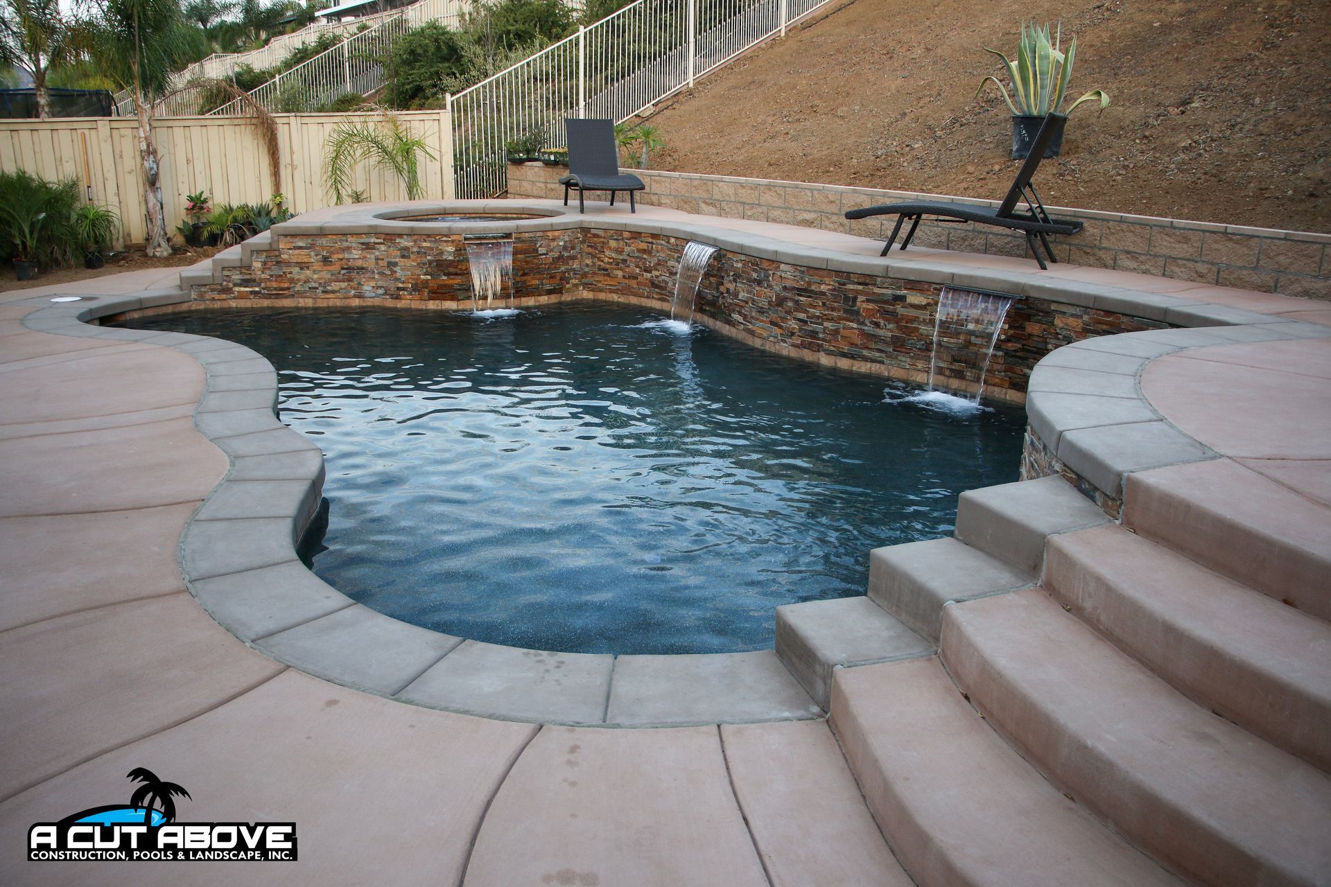 A freeform swimming pool with a stone wall water feature, sun loungers, and curved concrete steps in a backyard setting.
