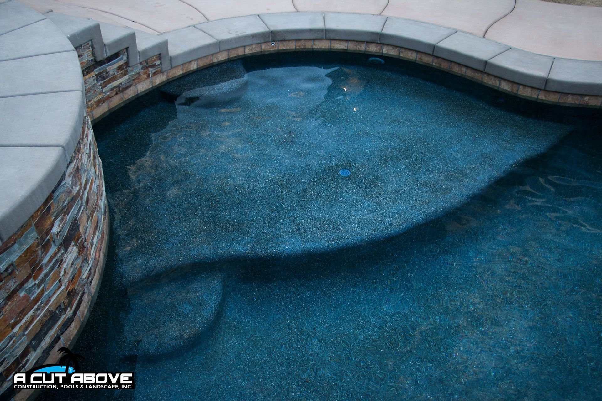 A high-angle view of a blue, pebble-finish pool with integrated steps, surrounded by stone coping and a paved deck.