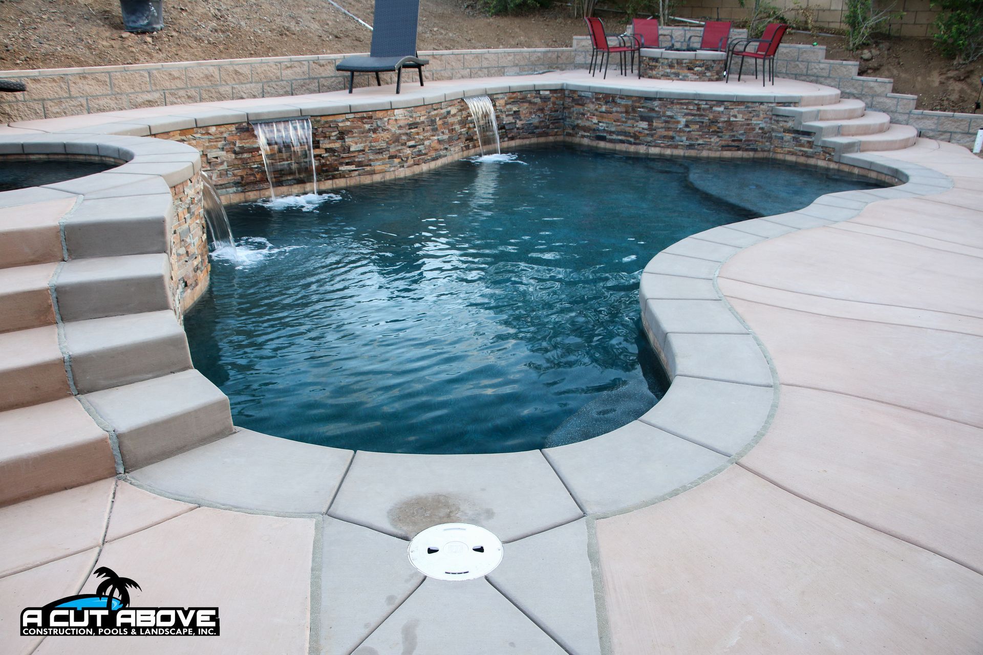 A backyard swimming pool with a stone water feature, a hot tub, and patio seating, surrounded by concrete decking.