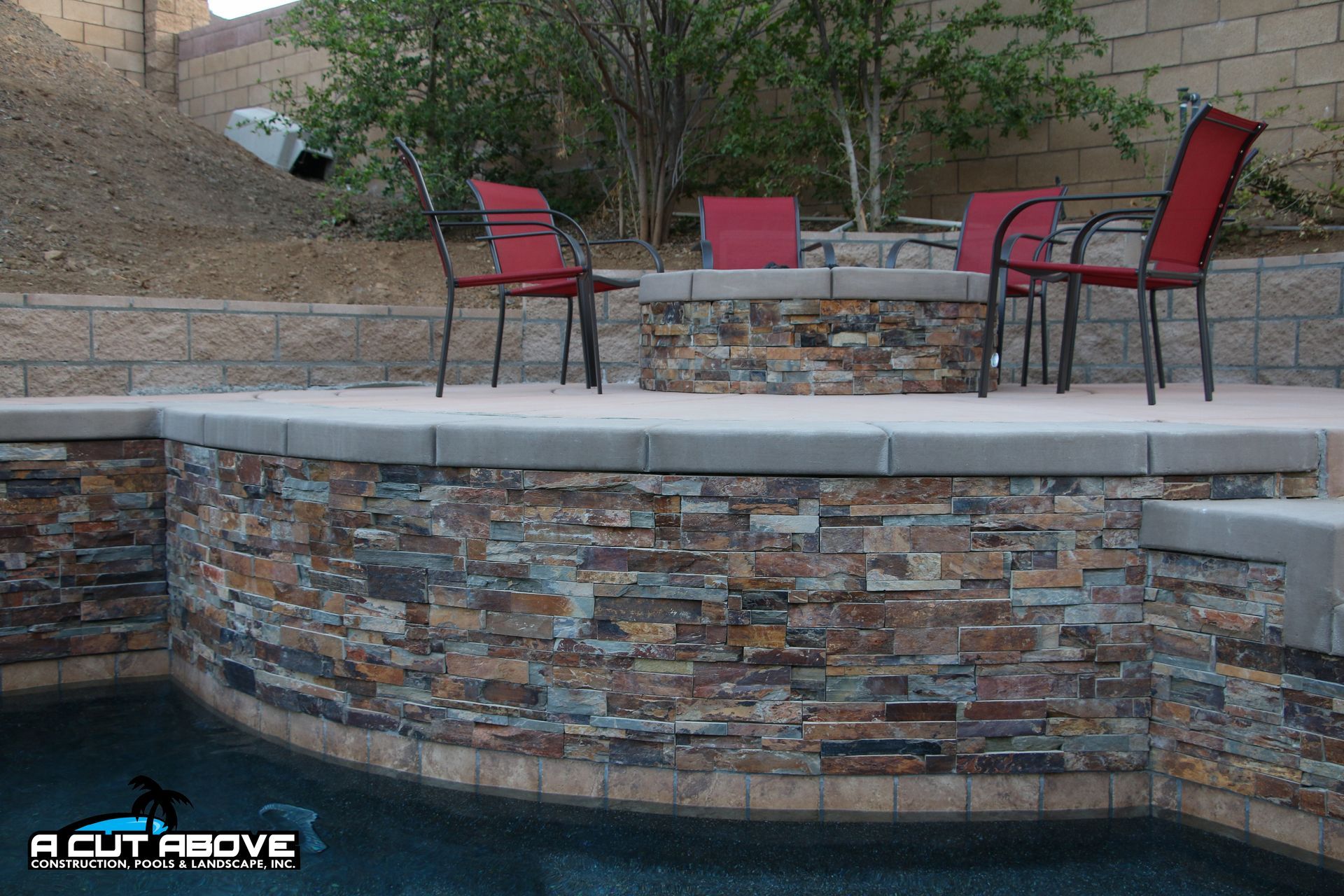 A tiered stone patio with a built-in fire pit and red chairs overlooking a swimming pool.