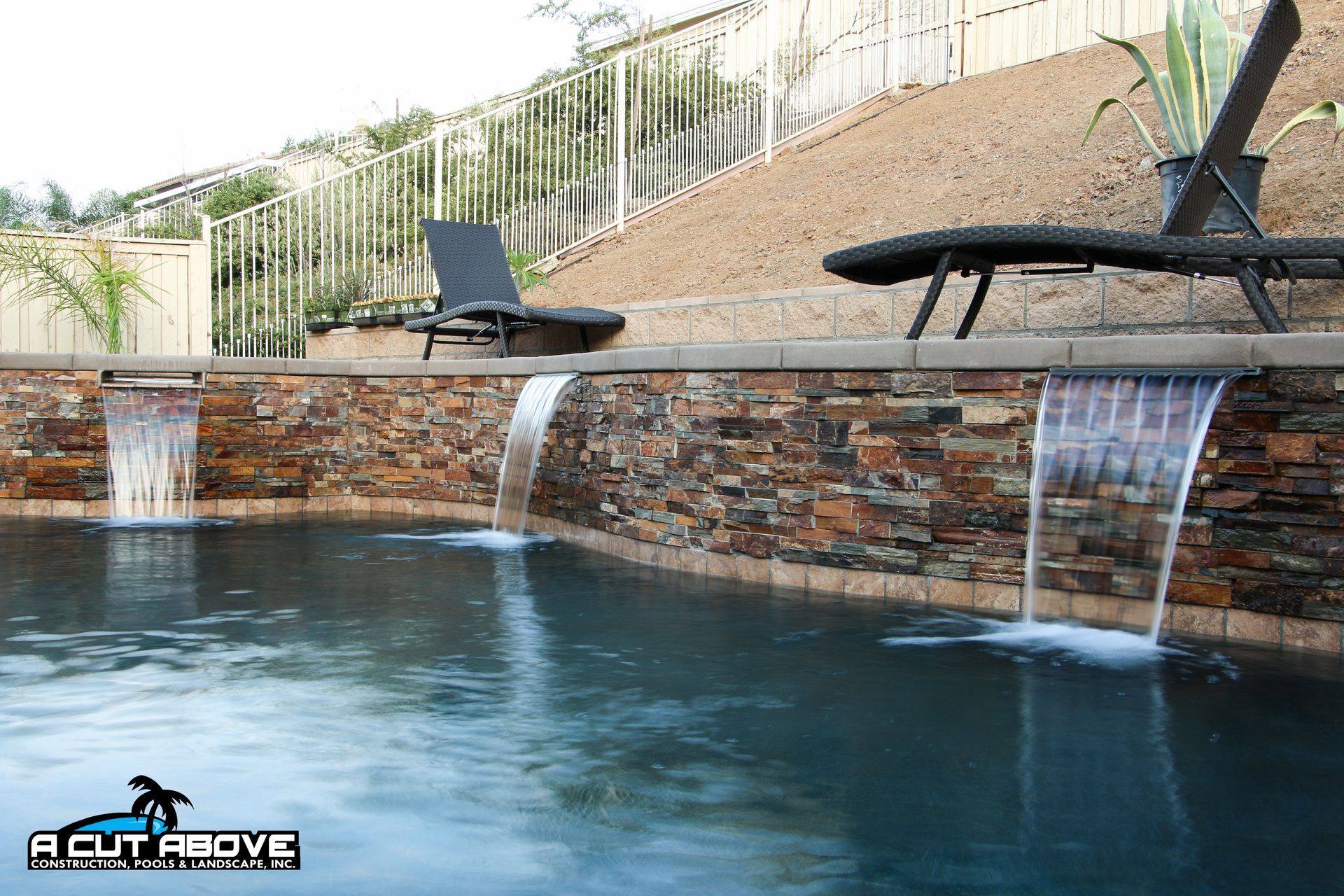 A stone-walled pool with three sheer descent waterfalls pouring into the dark water, beneath lounge chairs on a deck.