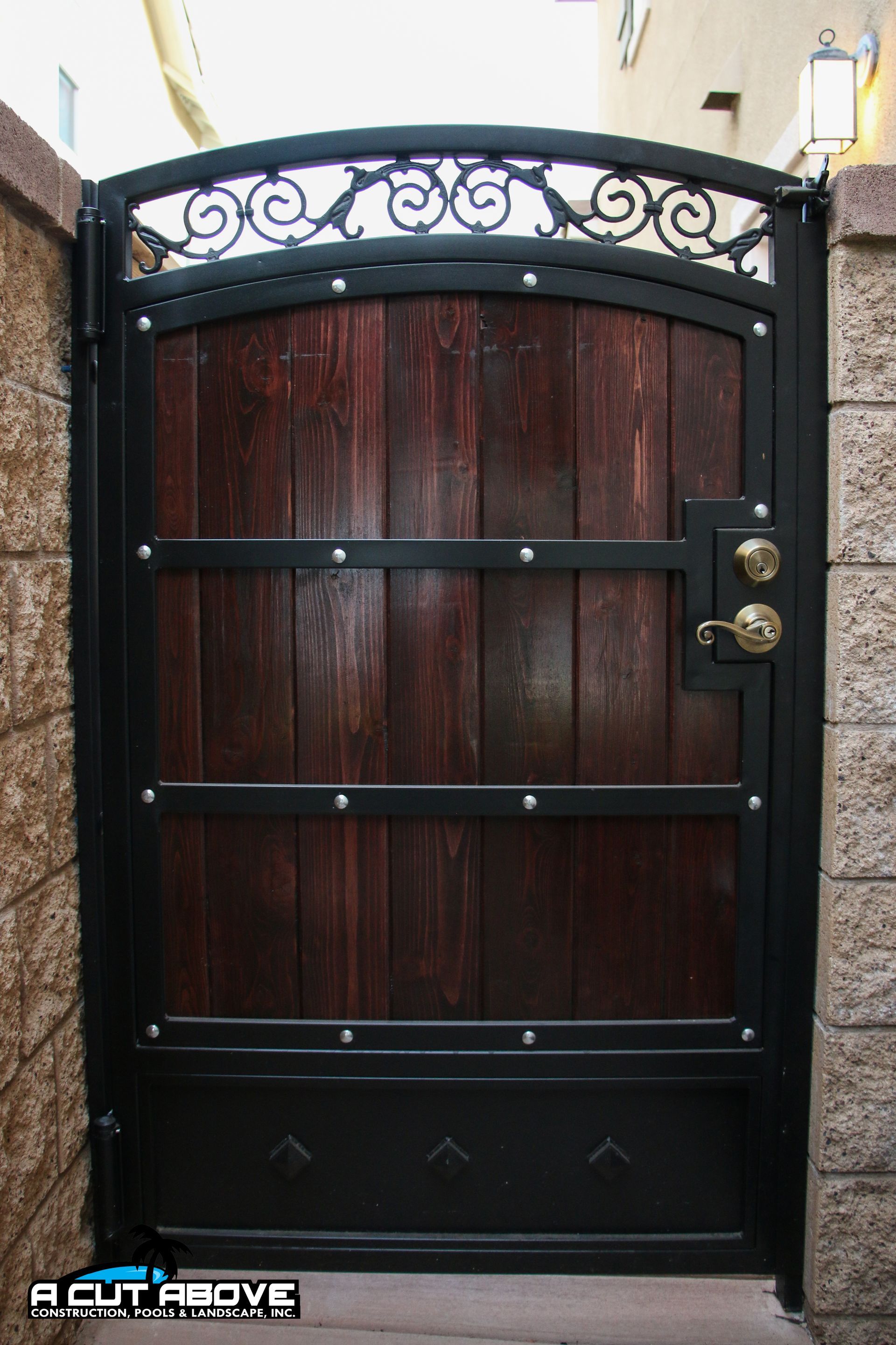 Arched gate with a dark wood panel center, black metal frame, scrollwork top, and metal hardware between stone walls.