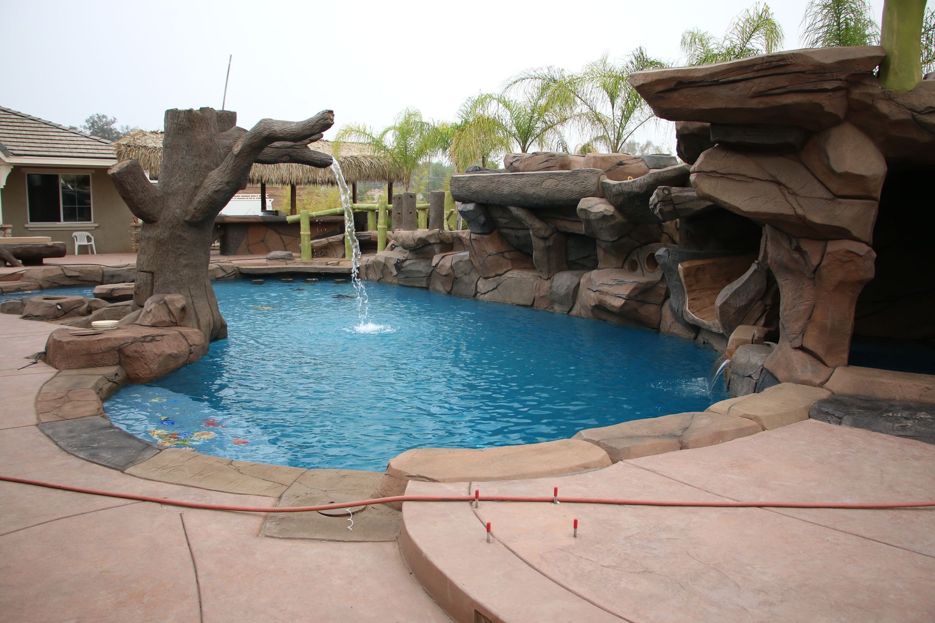 A backyard pool with a rock grotto, a waterslide, and a decorative tree water feature under a hazy sky.
