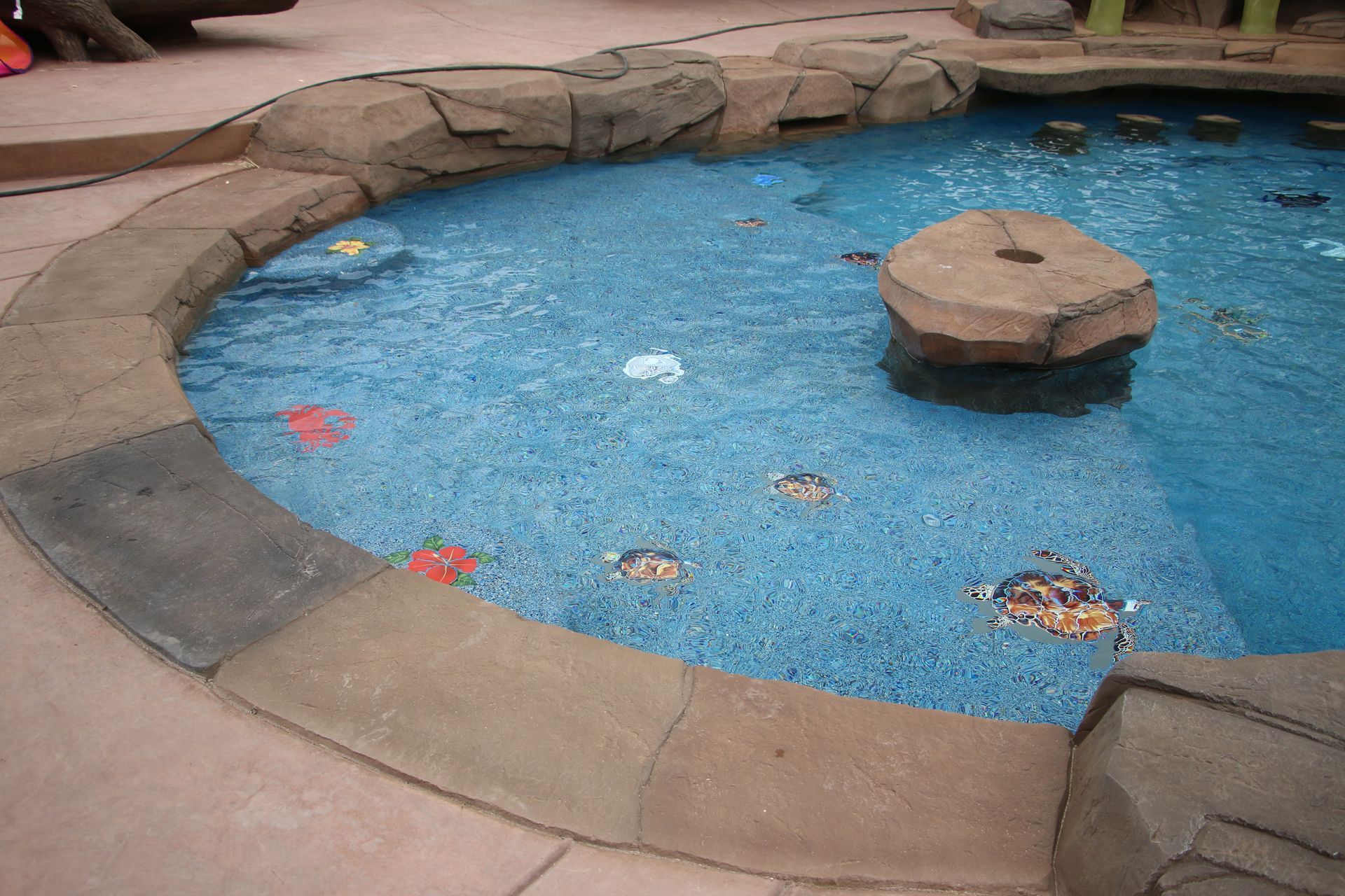 A small, blue-tiled circular wading pool with a stone island in the center and mosaic animal figures on the floor.