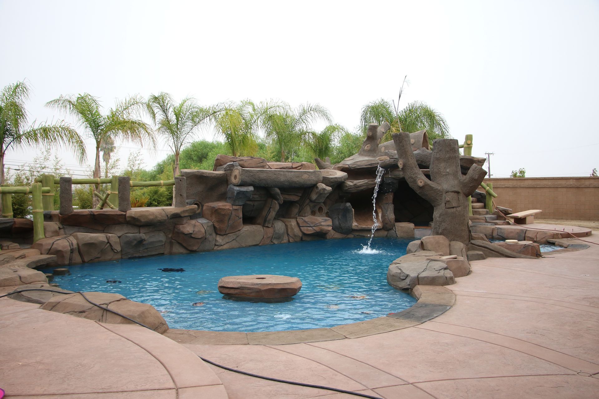 A decorative swimming pool with faux rock formations, a waterfall, and palm trees on a paved patio.