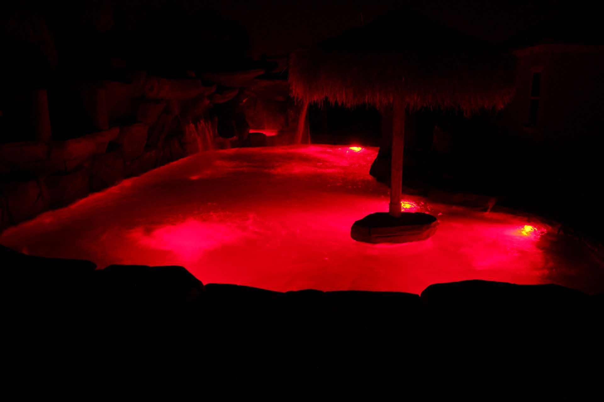 A swimming pool at night illuminated with vibrant red underwater lights, featuring a tiki-style umbrella stand.