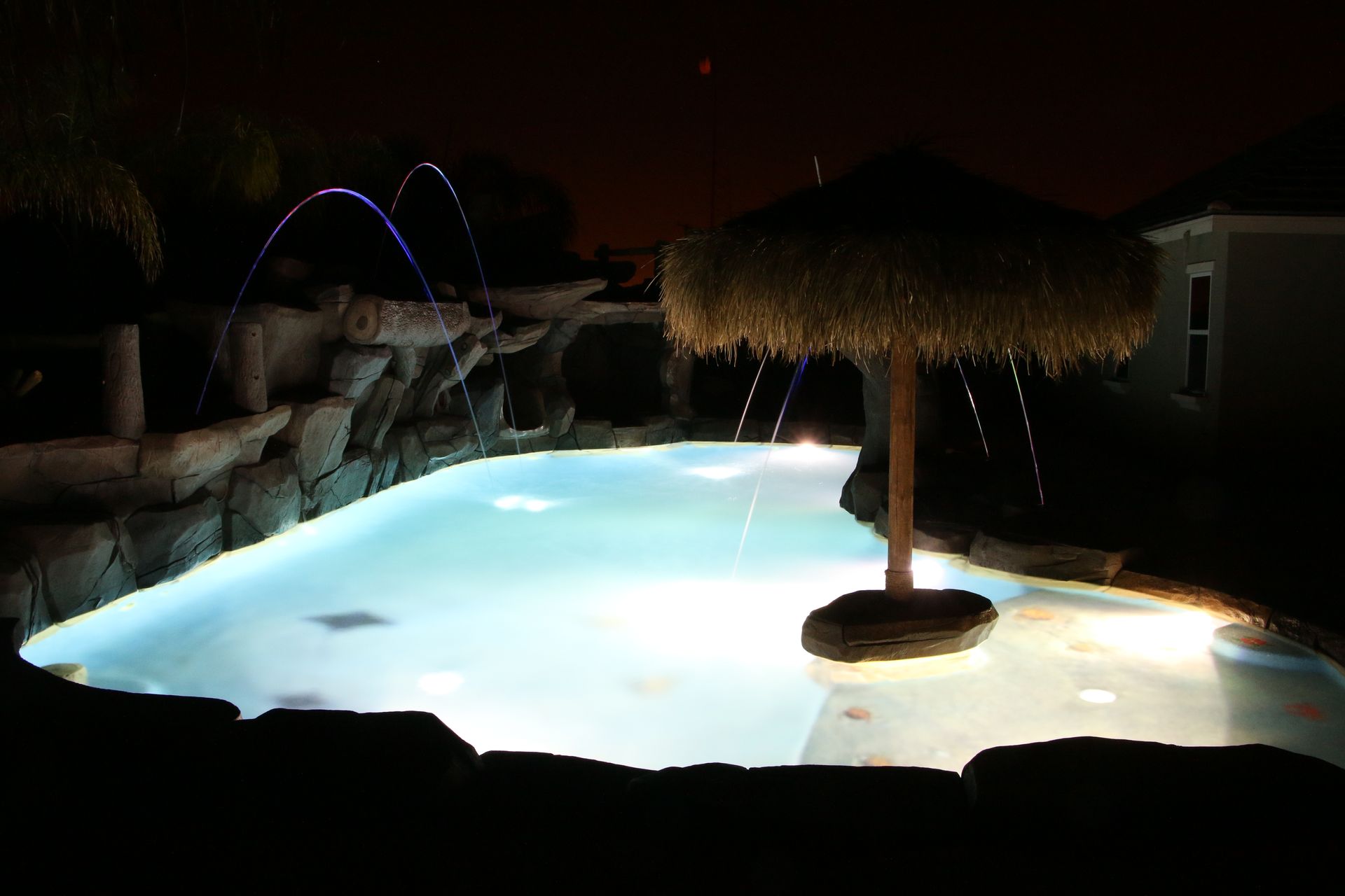 A brightly lit swimming pool at night featuring a tiki-style umbrella, rock water features, and arching water jets.