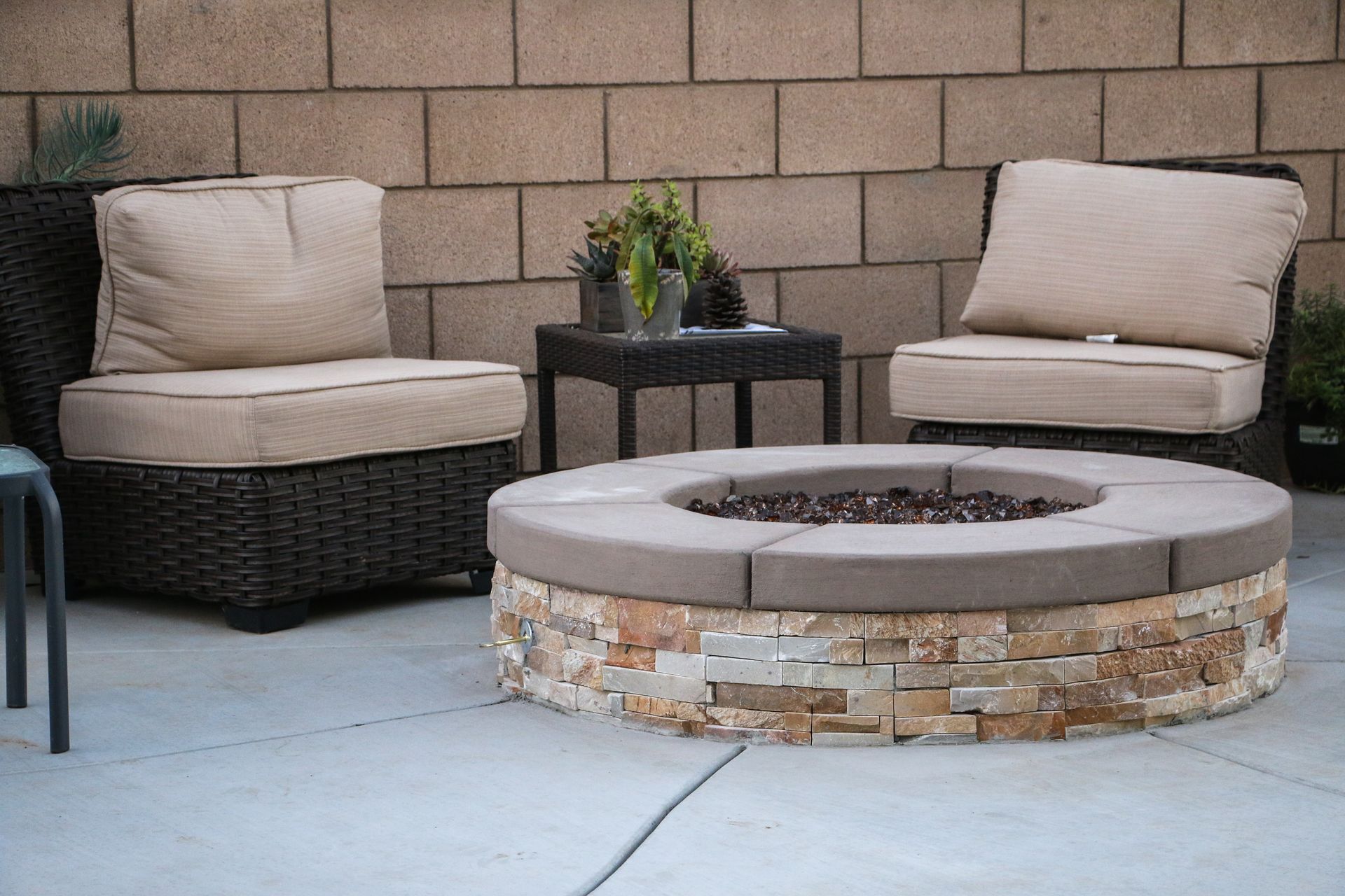 Two wicker armchairs with tan cushions and a small table flank a round, stacked-stone fire pit on a concrete patio.
