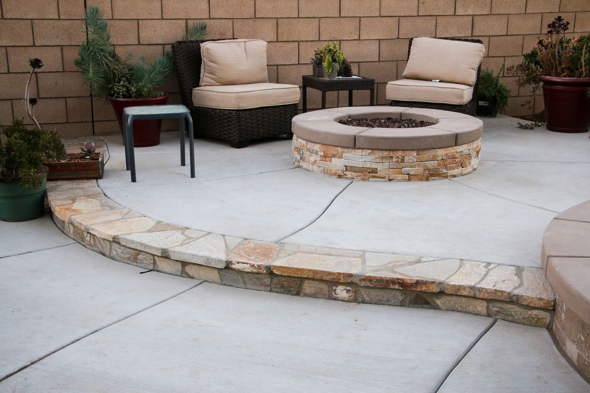 A patio with a stone fire pit, two wicker chairs, a small table, and potted plants against a concrete block wall.