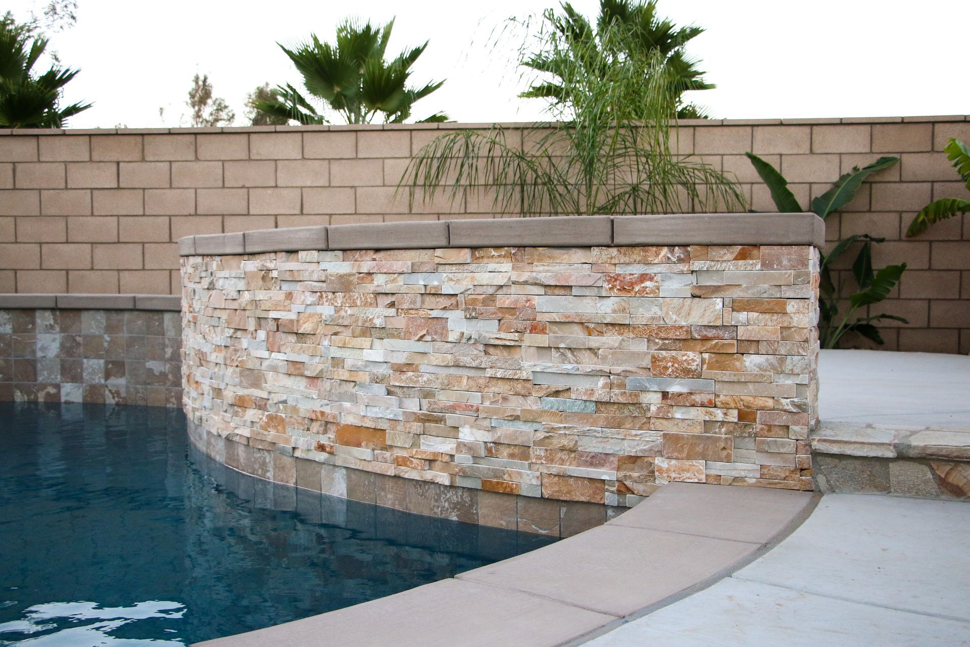 Curved stone wall bordering a swimming pool, set against a tan brick wall and palm plants in a backyard patio area.