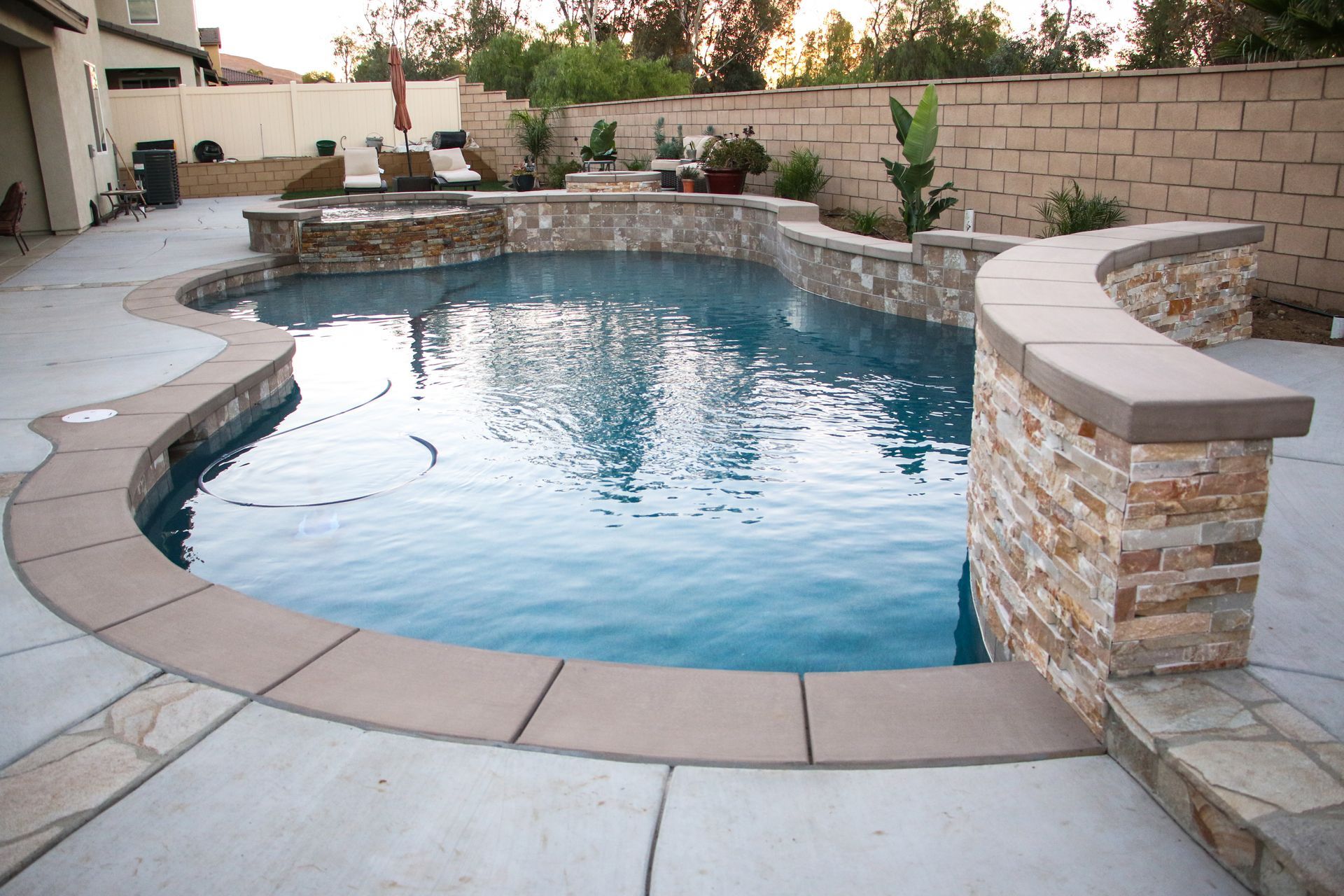 A backyard swimming pool with blue water, surrounded by stone-clad retaining walls and beige concrete decking.