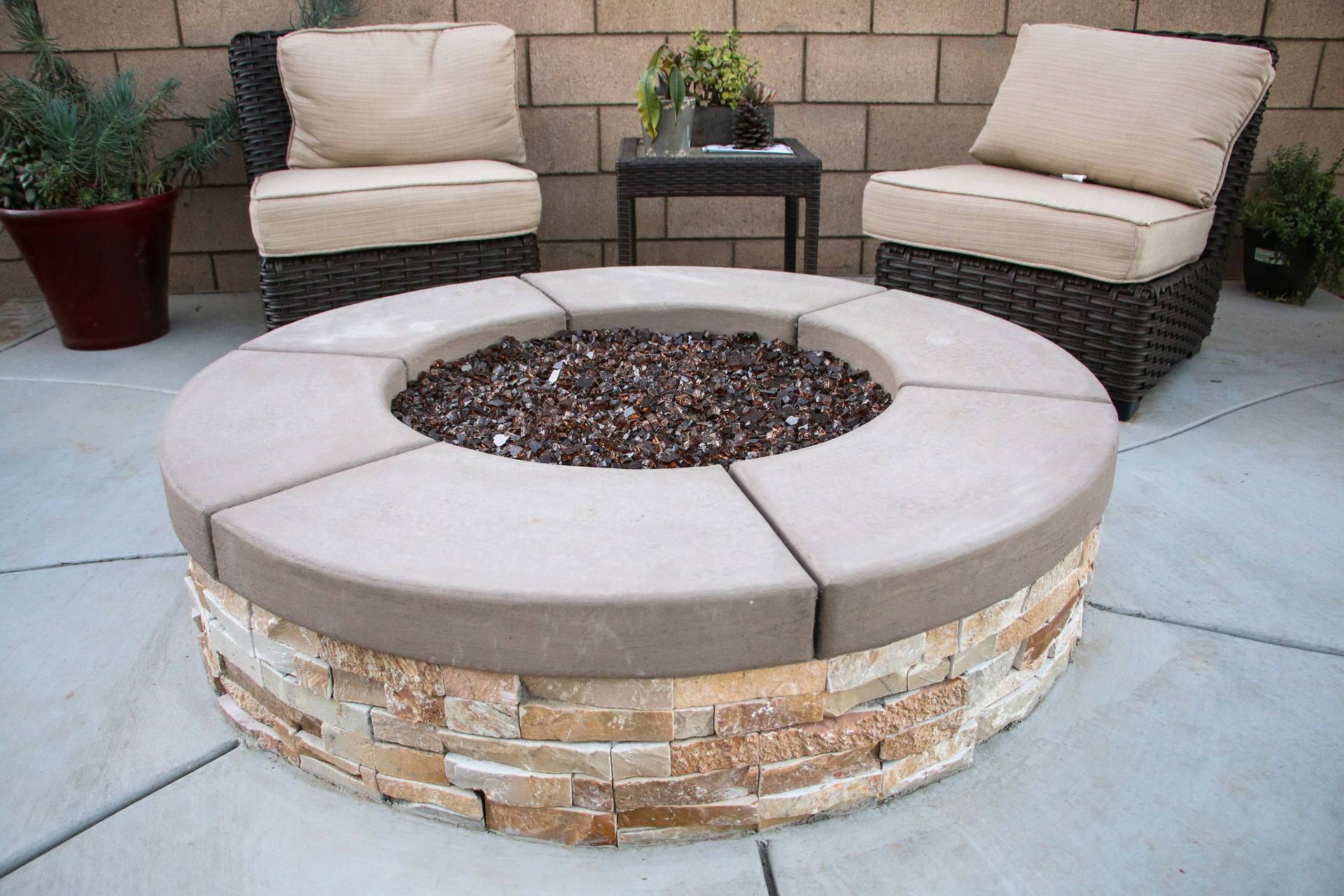 Round stone fire pit with a gravel center, surrounded by two wicker chairs and a small table on a concrete patio.