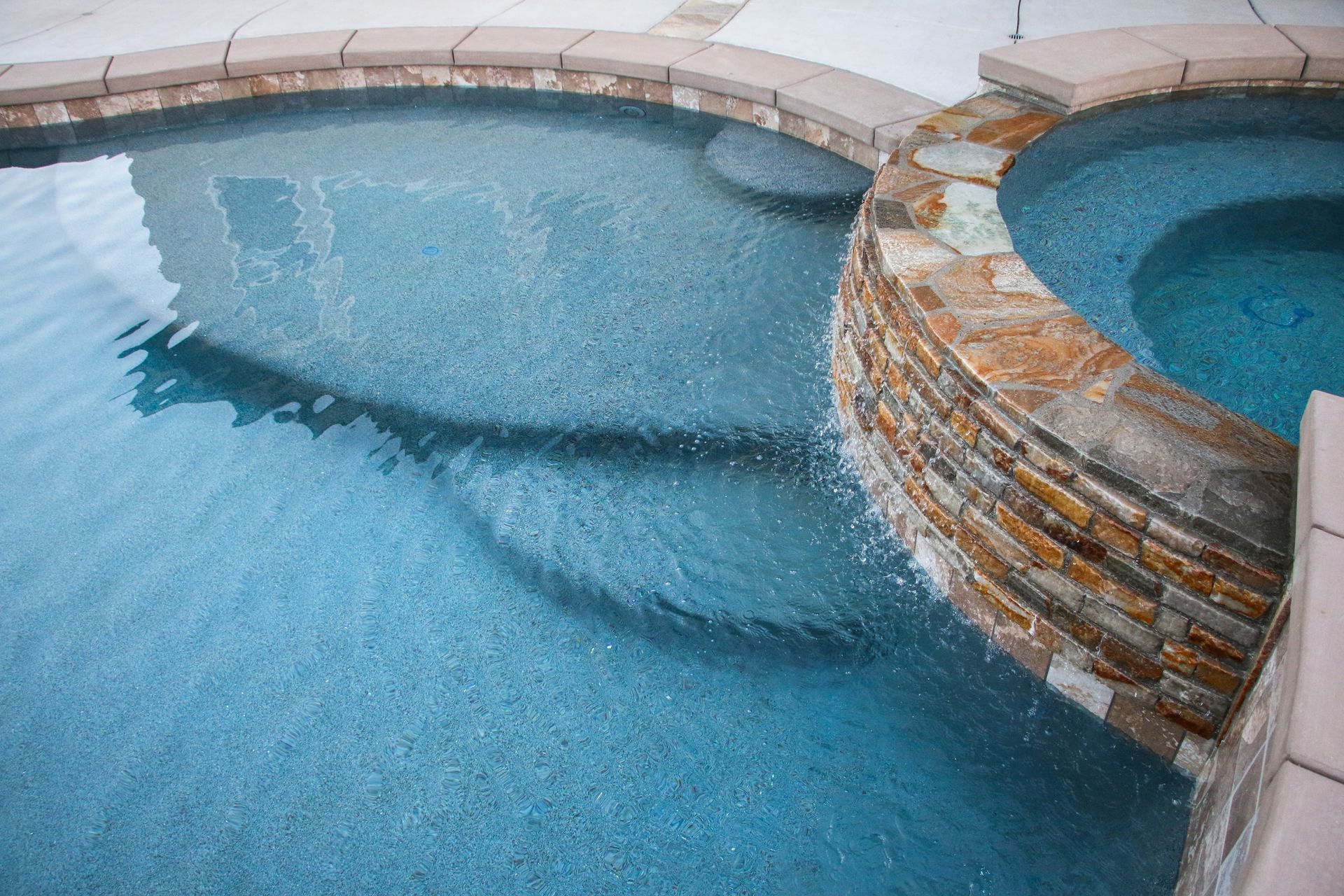 A stone-walled spa spilling water into a clear, blue swimming pool with curved steps.