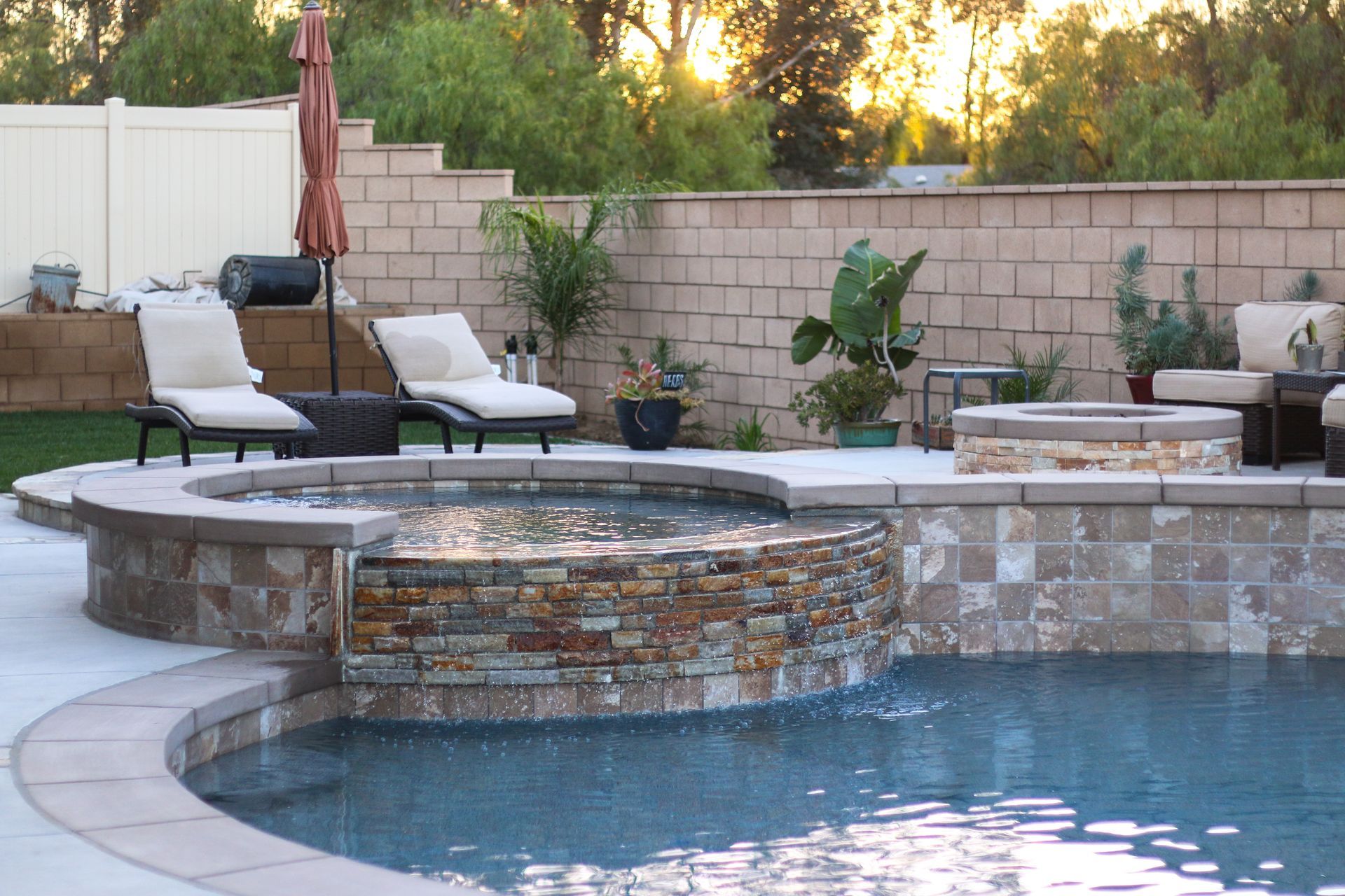 A backyard swimming pool with a stone hot tub spillover, lounge chairs, a fire pit, and potted plants at sunset.
