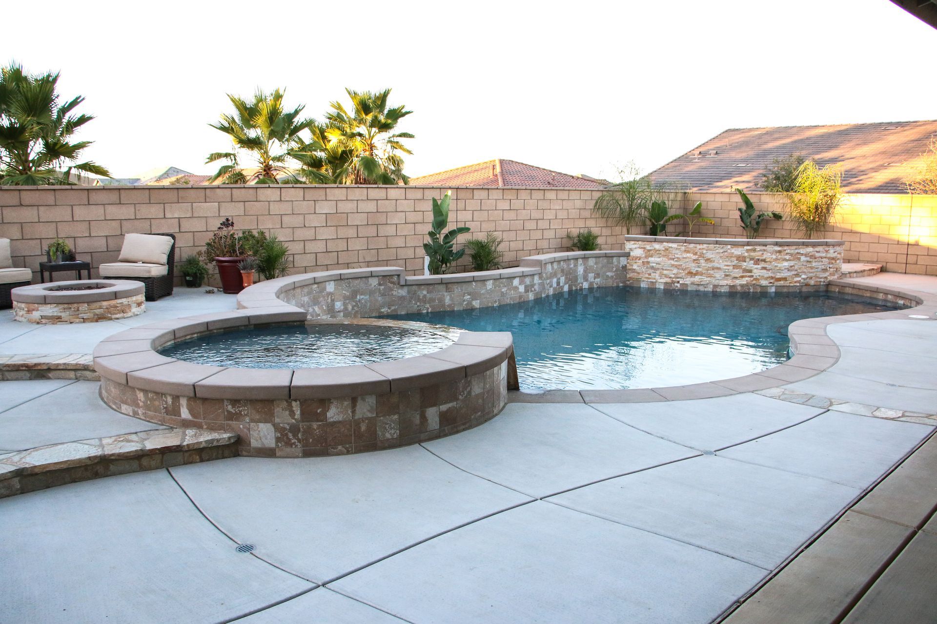 A backyard swimming pool with an integrated spa, stone walls, and a fire pit, set against a tan wall and hills.