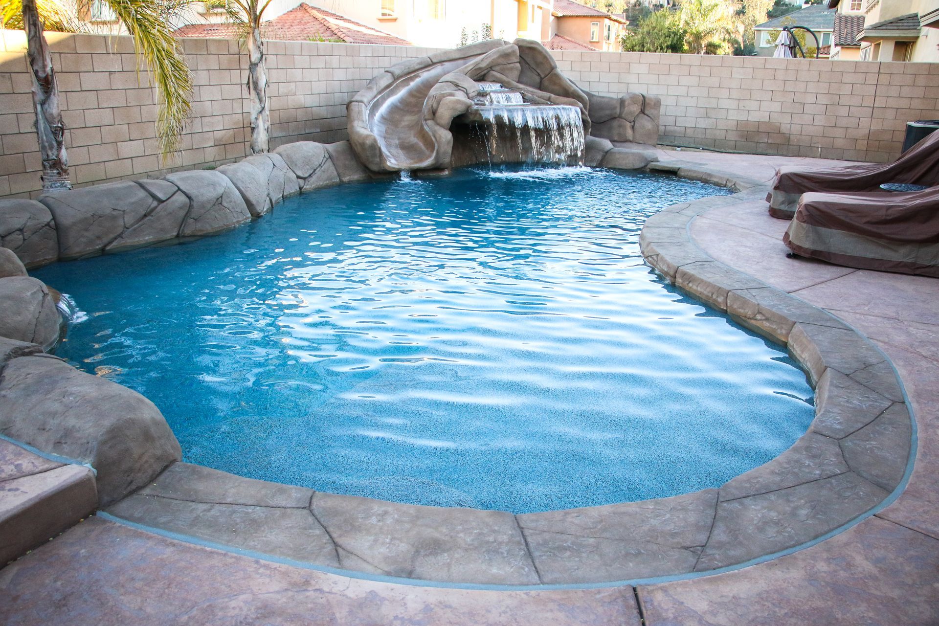 A swimming pool with a rock-style slide and waterfall feature, surrounded by tan stone coping and a paved patio.