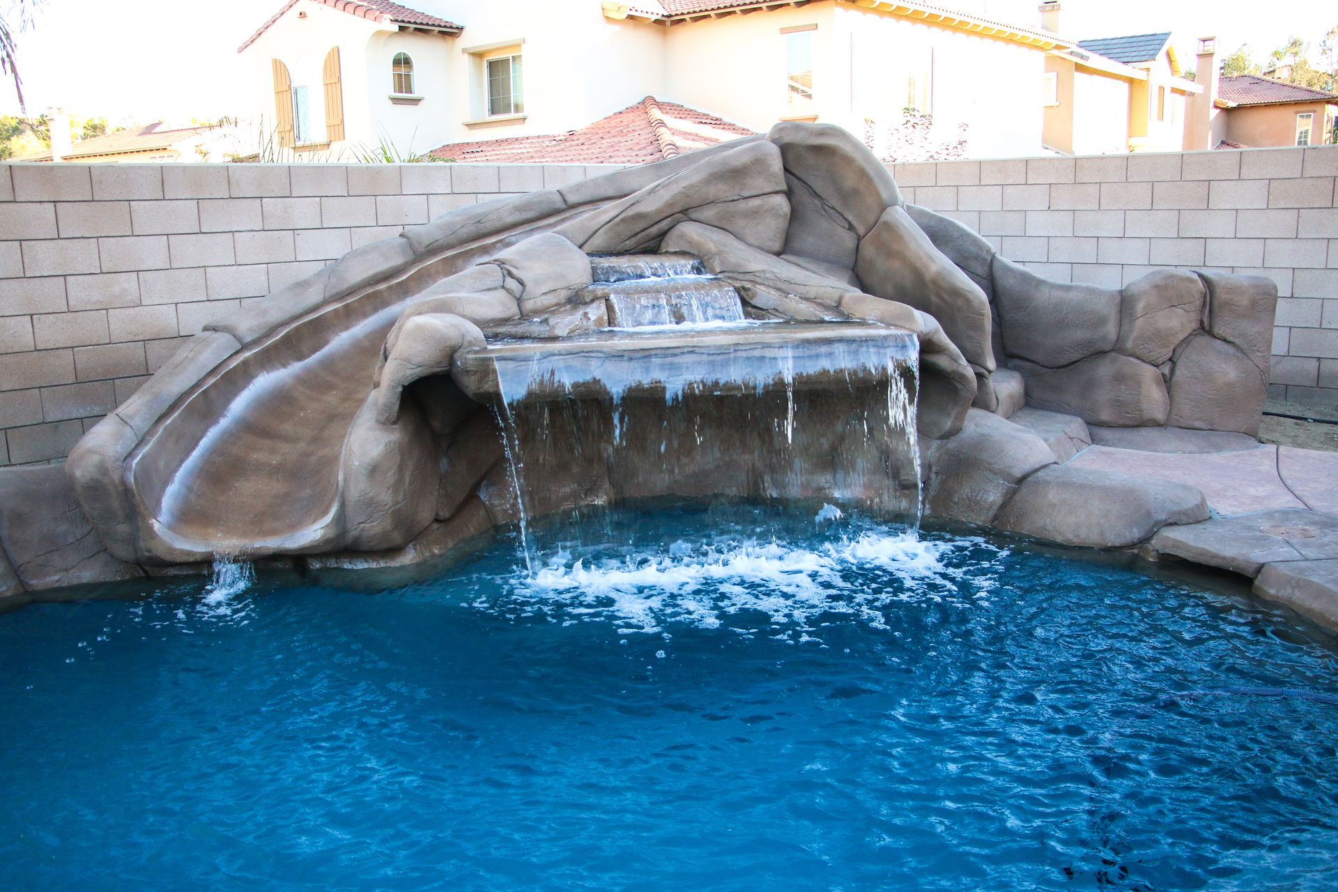 A brown rock-style pool water slide and waterfall feature spilling into a blue swimming pool in a backyard.