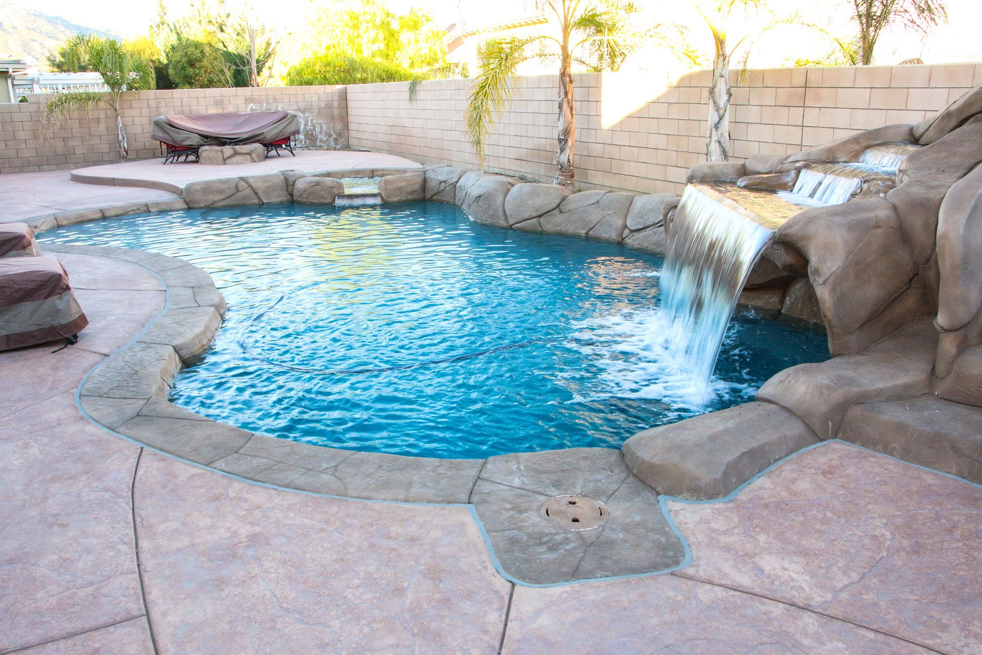 A curved backyard swimming pool with a stone rock waterfall feature, concrete decking, and a stone privacy wall.