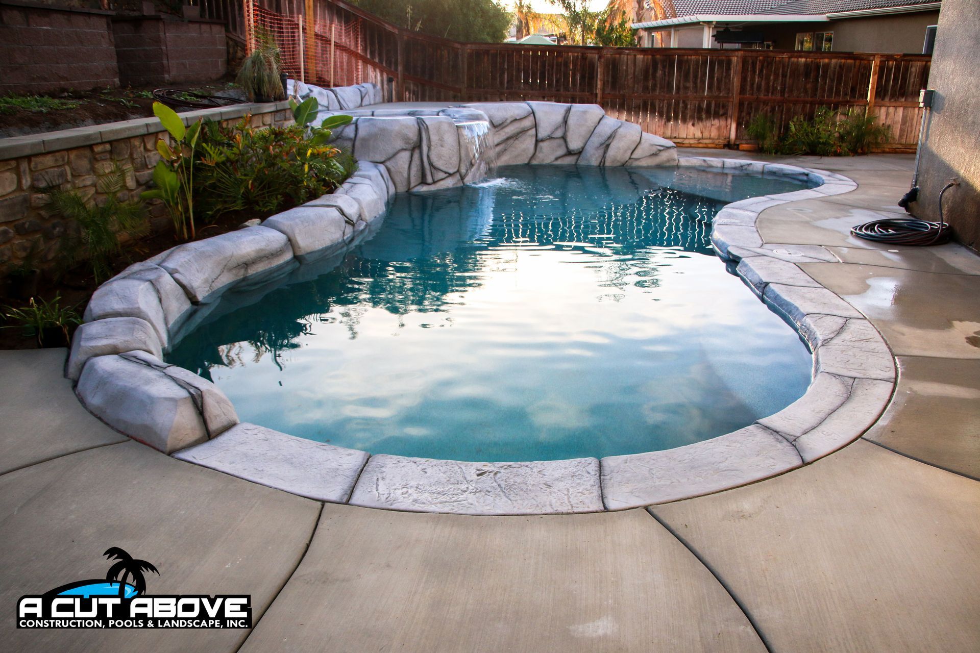 A backyard swimming pool with custom faux-rock features, a water feature, and a paved deck, viewed in warm lighting.