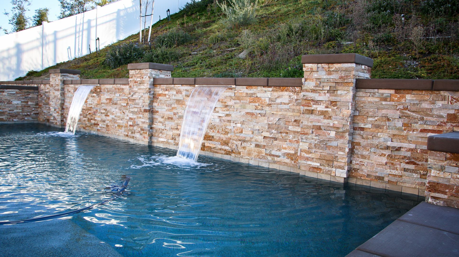 A backyard swimming pool features a tan stone wall with two water fountains spilling into the blue water.