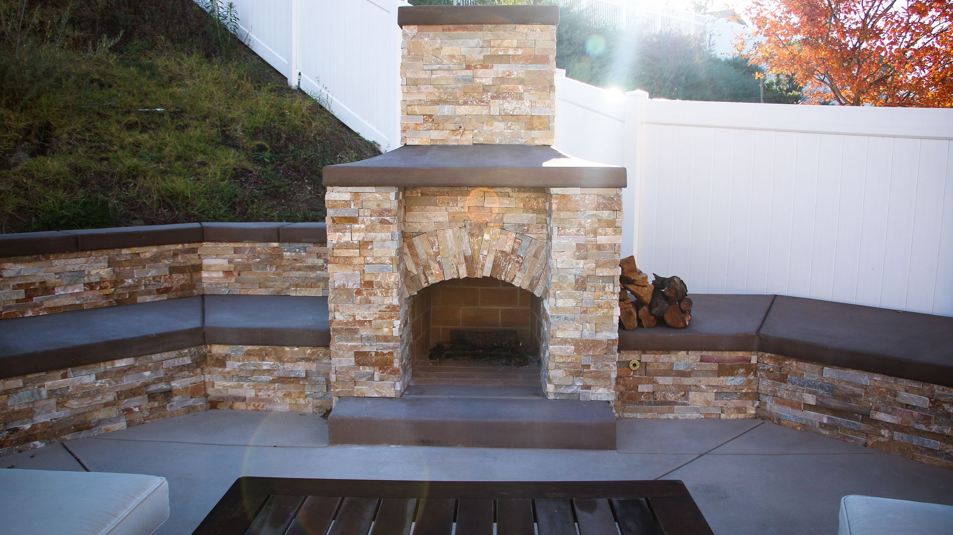 A stone outdoor fireplace with built-in curved seating and dark concrete counters, set against a white fence in a yard.