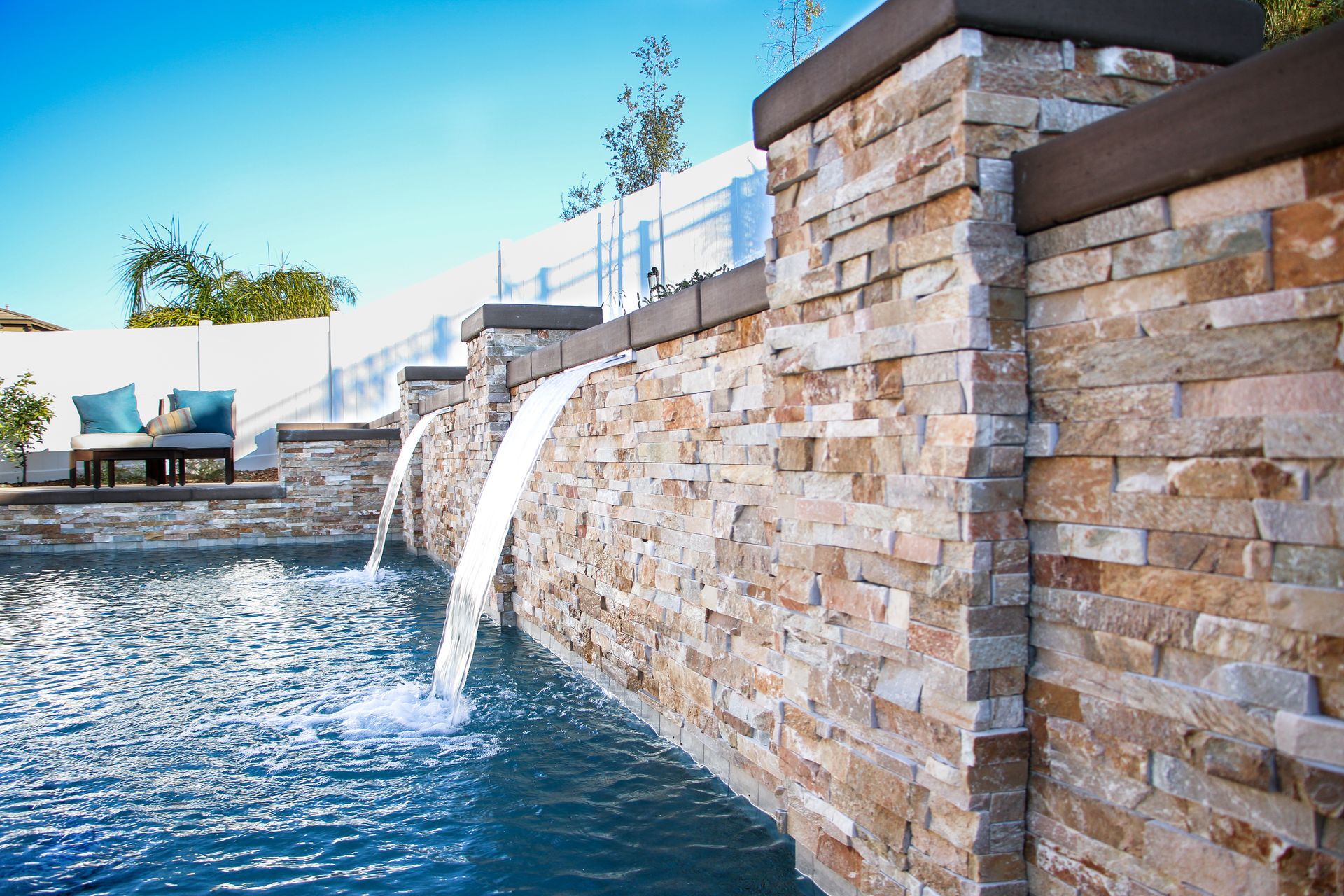 Stone water feature with two cascading waterfalls flowing into a swimming pool on a sunny day.
