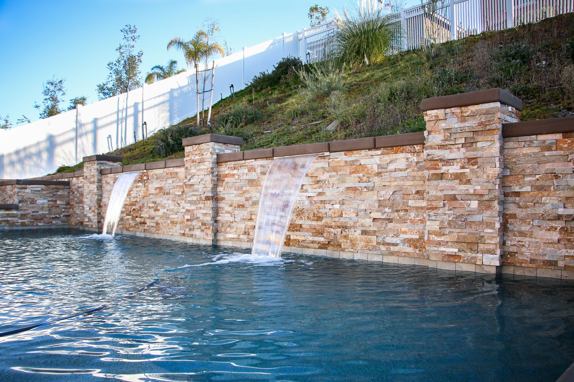 A stone retaining wall with two water features flowing into a swimming pool below, set against a hillside with greenery.