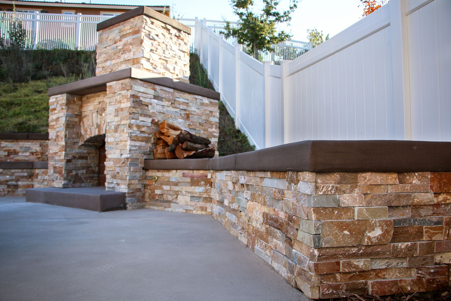 Outdoor stone fireplace and seating wall on a concrete patio next to a white fence in a backyard setting.