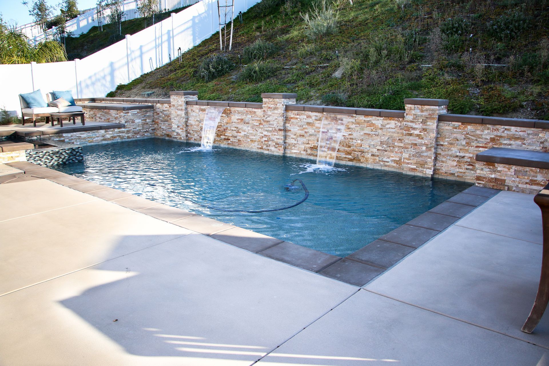 A rectangular swimming pool with stone waterfall features, bordered by a concrete patio and a sloped, landscaped hill.