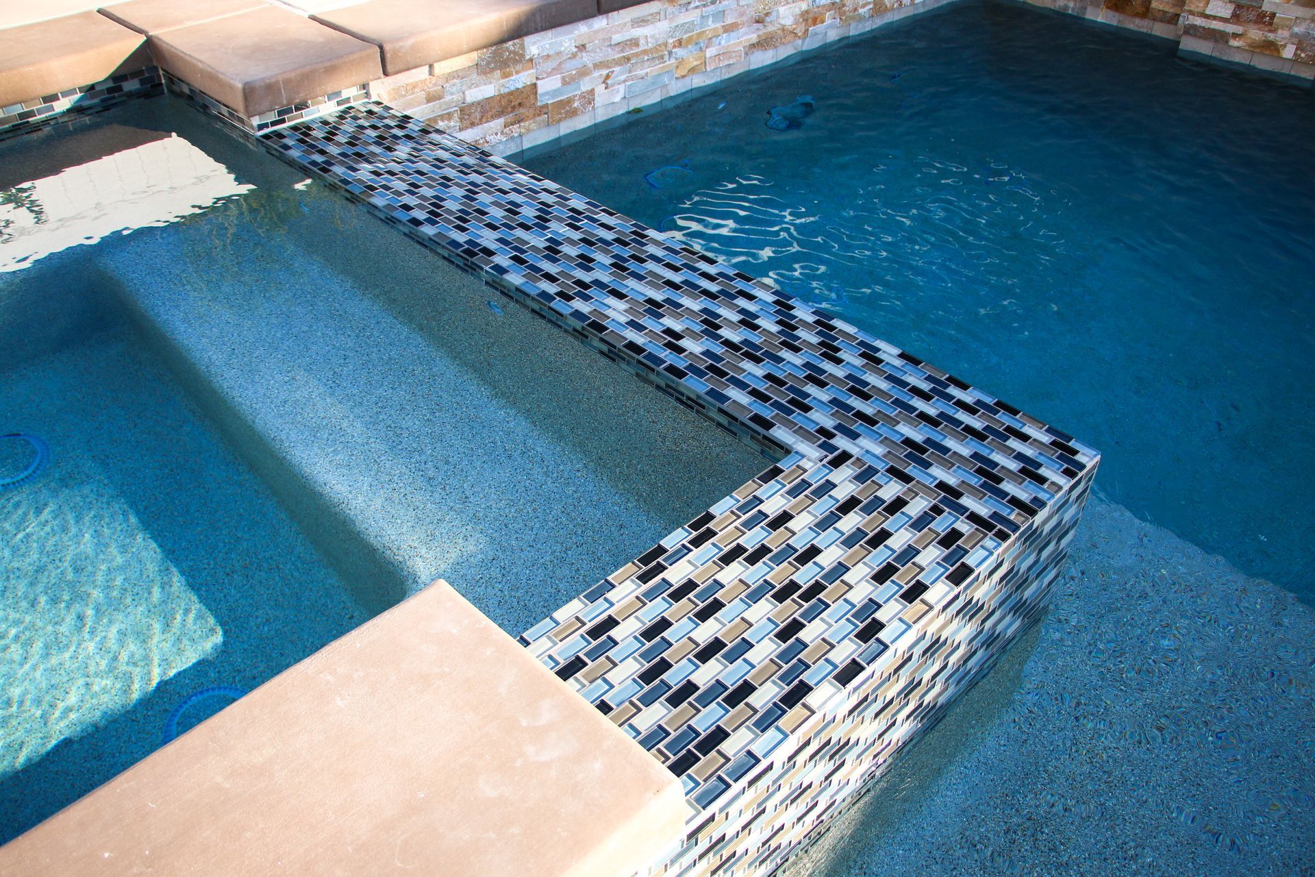 An L-shaped spa spillway with multi-colored glass tiles, separating it from the main swimming pool.