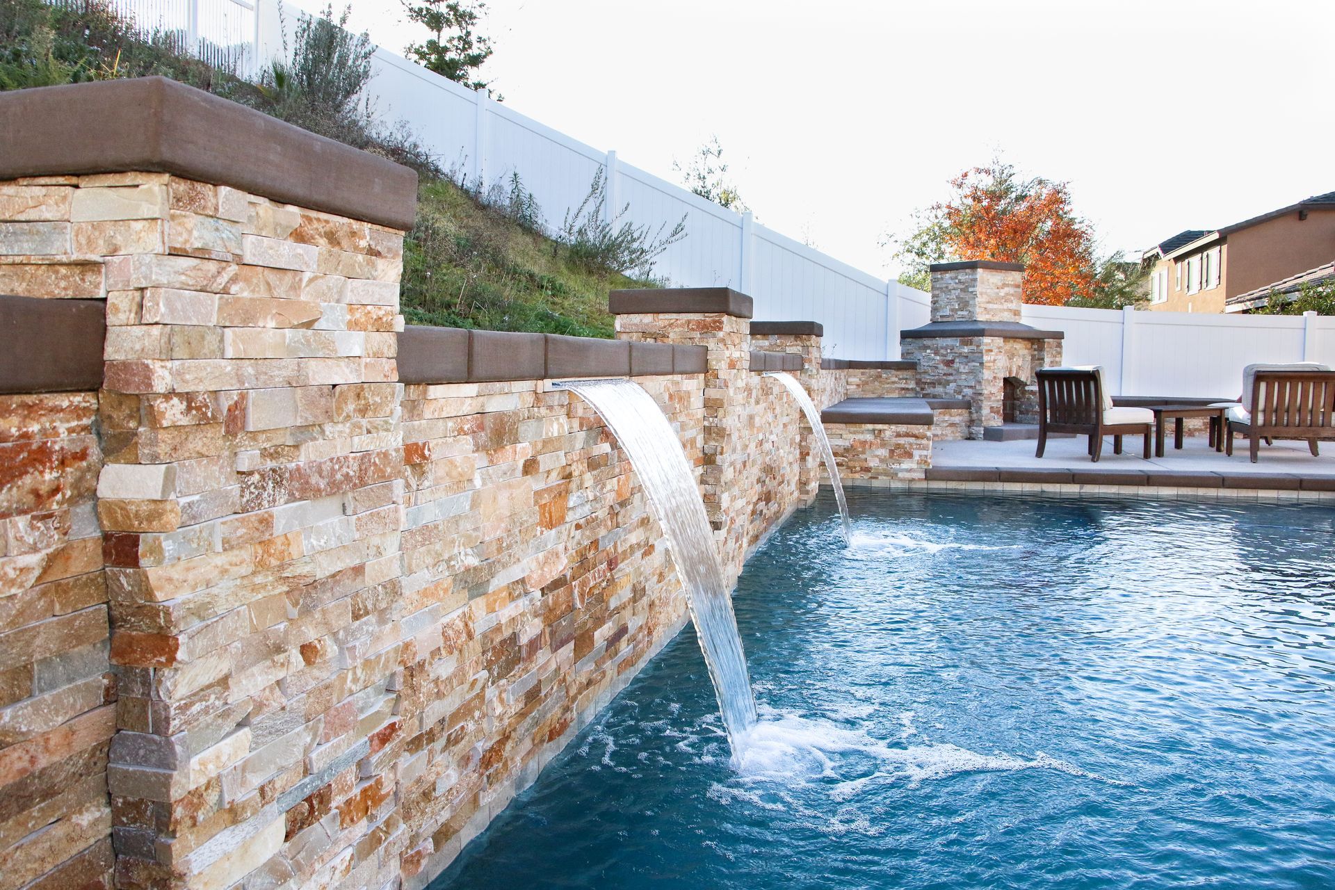 A stone water feature with two fountains cascading into a pool in a backyard setting with patio furniture.