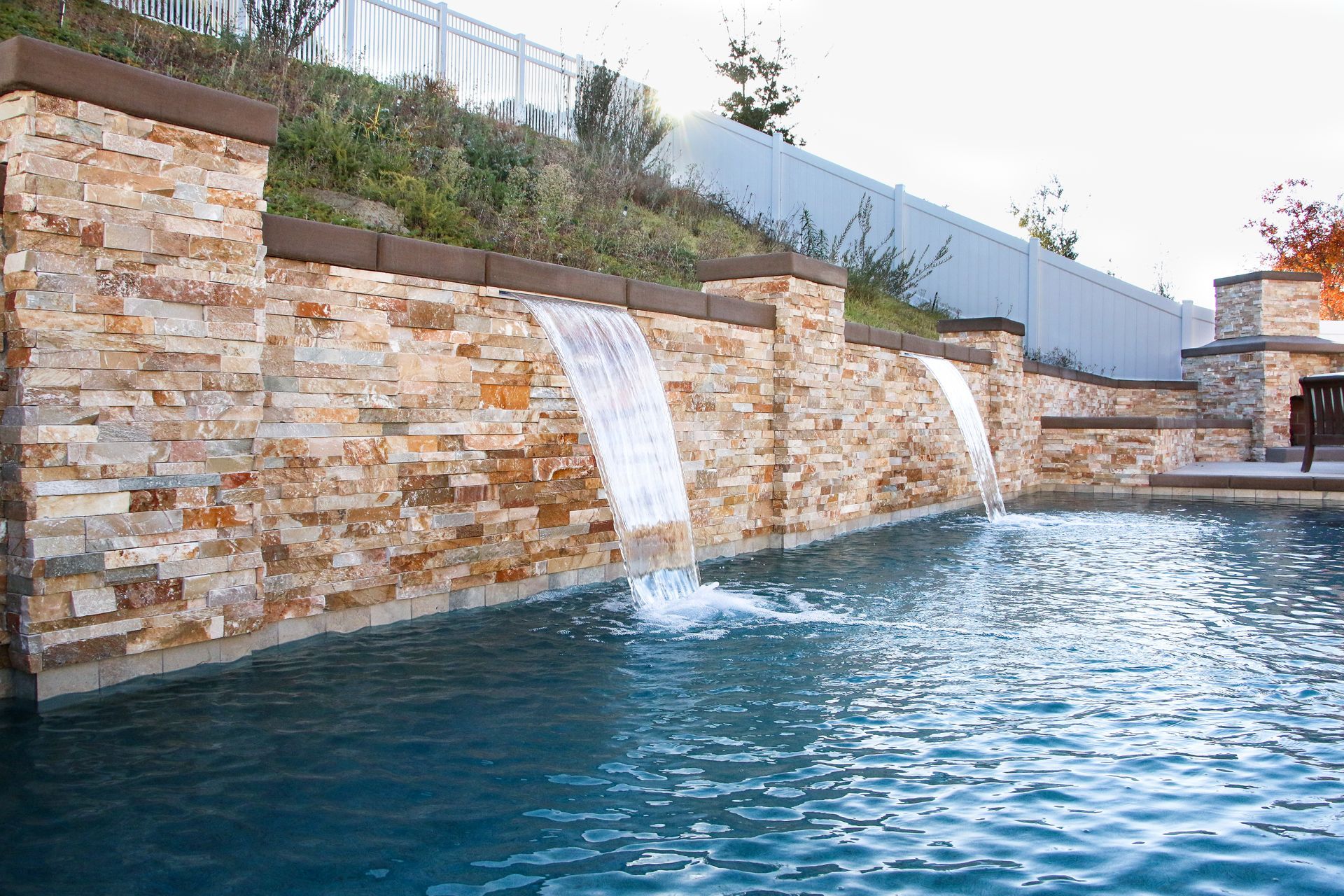 A stone wall with two water features flowing into a backyard swimming pool at twilight.