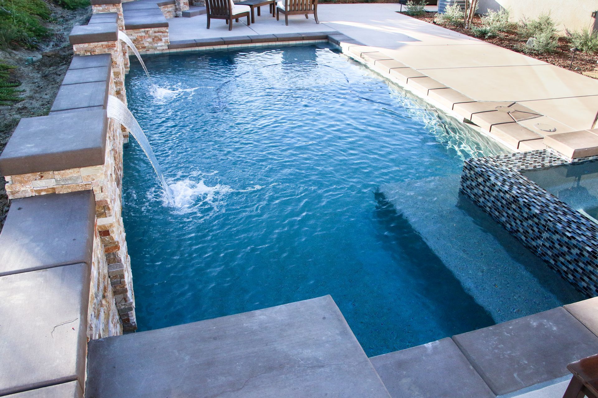 A rectangular backyard pool with a stone wall water feature and a stone-tiled sitting area nearby.