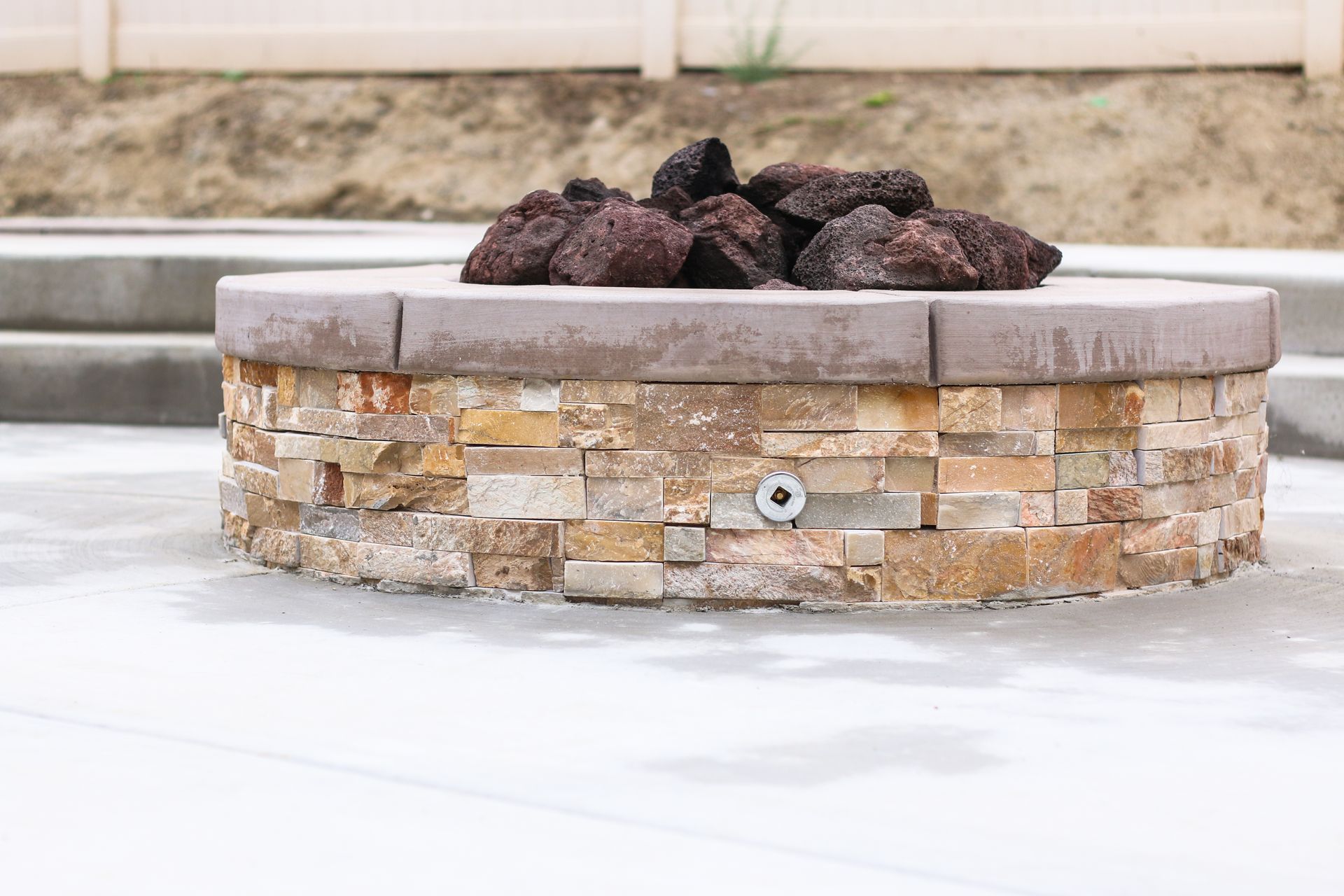 A circular stone fire pit with dark lava rocks on top, set on a light-colored patio.