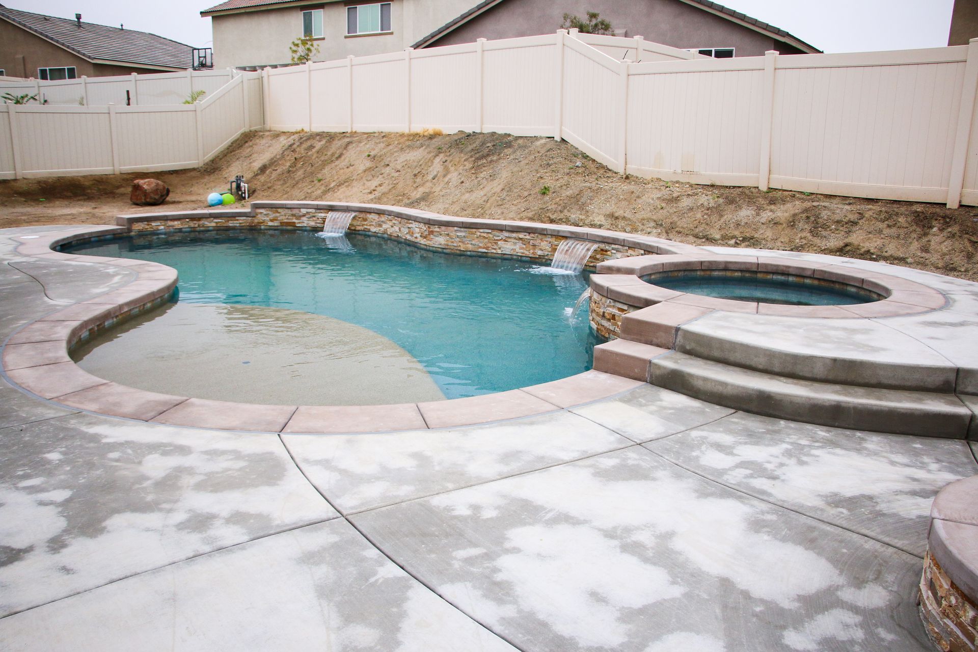 A backyard swimming pool with a connected circular spa, tanning ledge, and water features, surrounded by concrete decking.