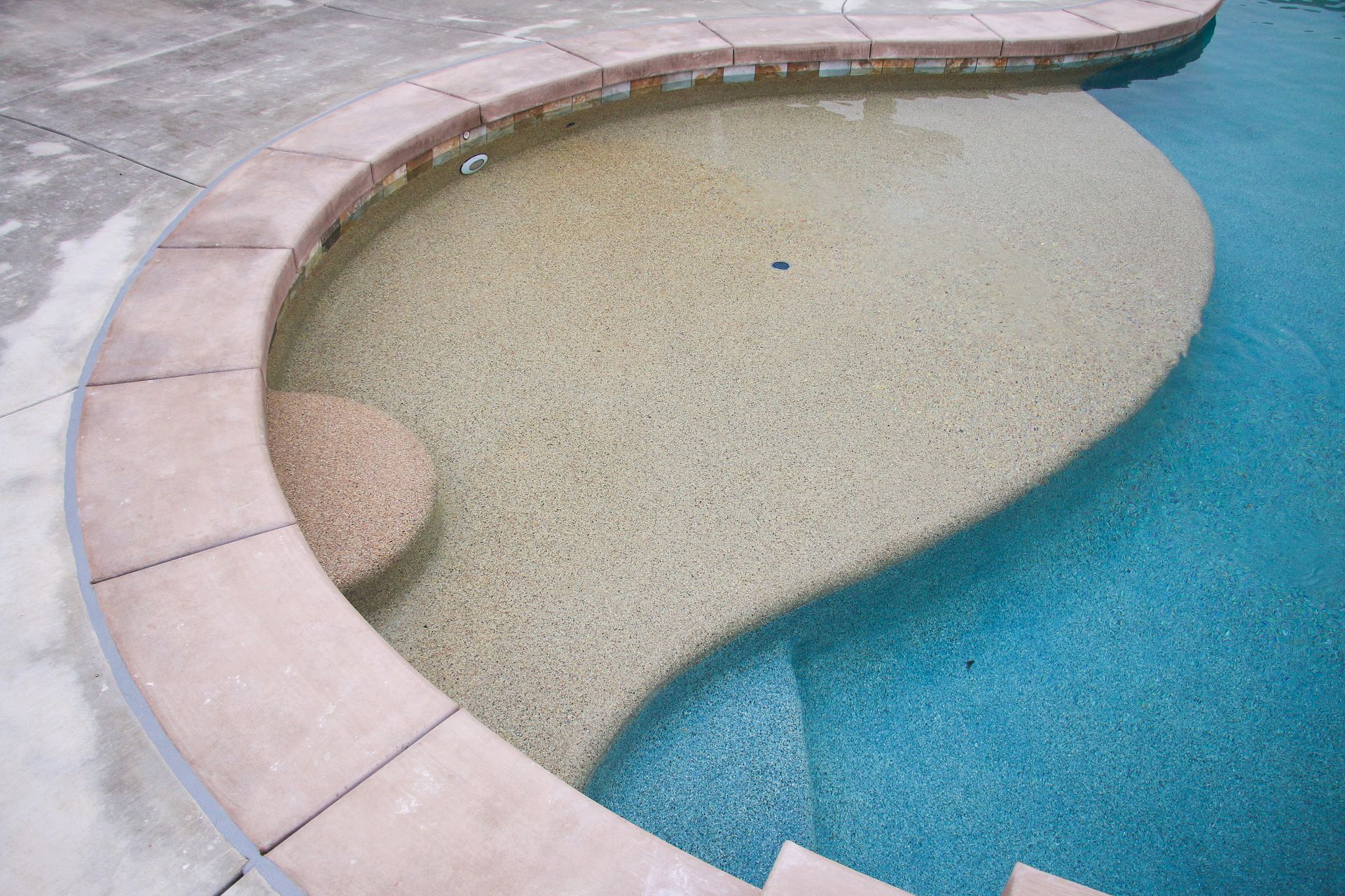 A wide, curved pool tanning ledge with light-colored pebble finish, bordered by concrete coping at the edge of a blue pool.