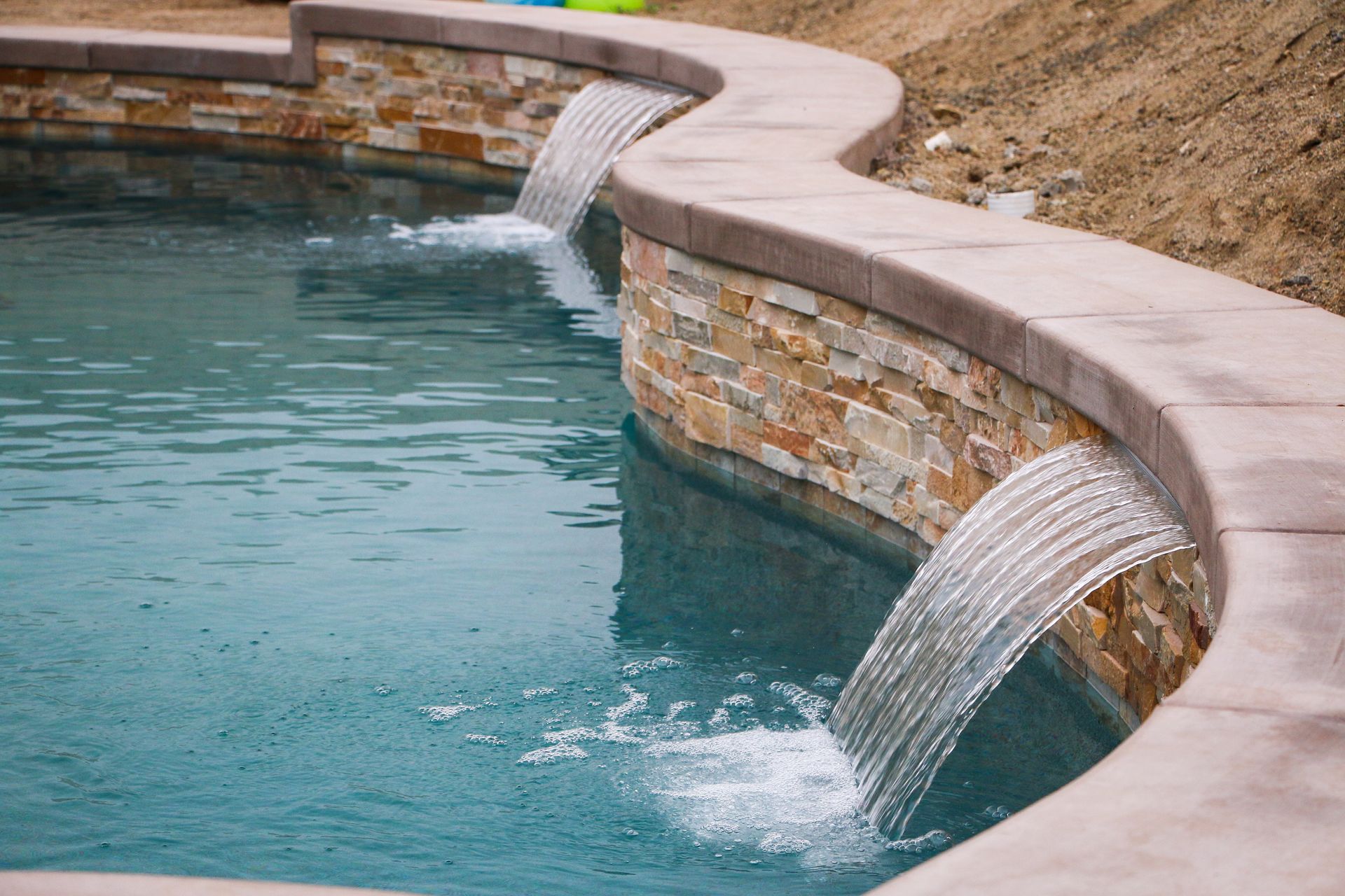Two cascading water features flowing from a curved, stone-tiled wall into a swimming pool.