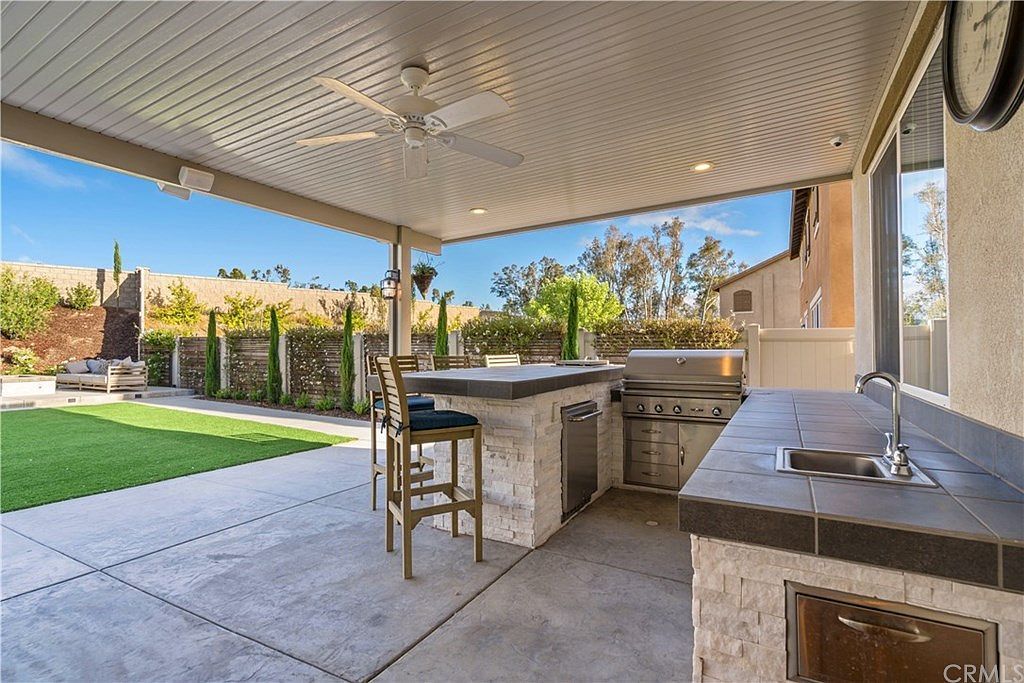 Outdoor kitchen on a covered patio with tile countertops, a sink, a grill, and a tall chair overlooking a green lawn.