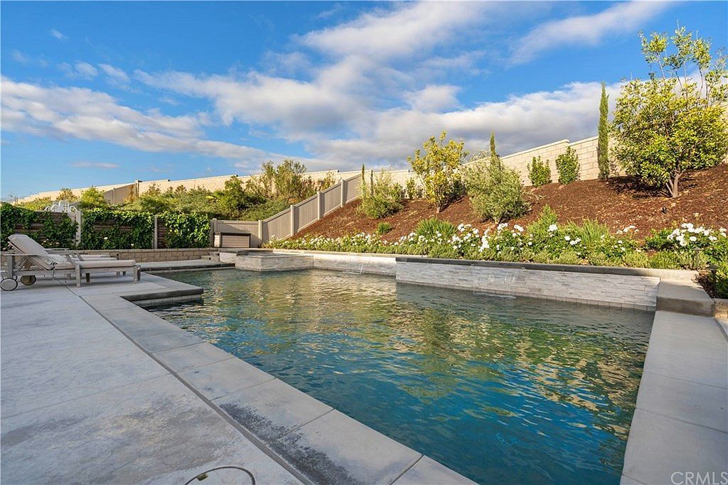 A swimming pool with stone accents, surrounded by landscaping and a stone wall under a blue sky with clouds.
