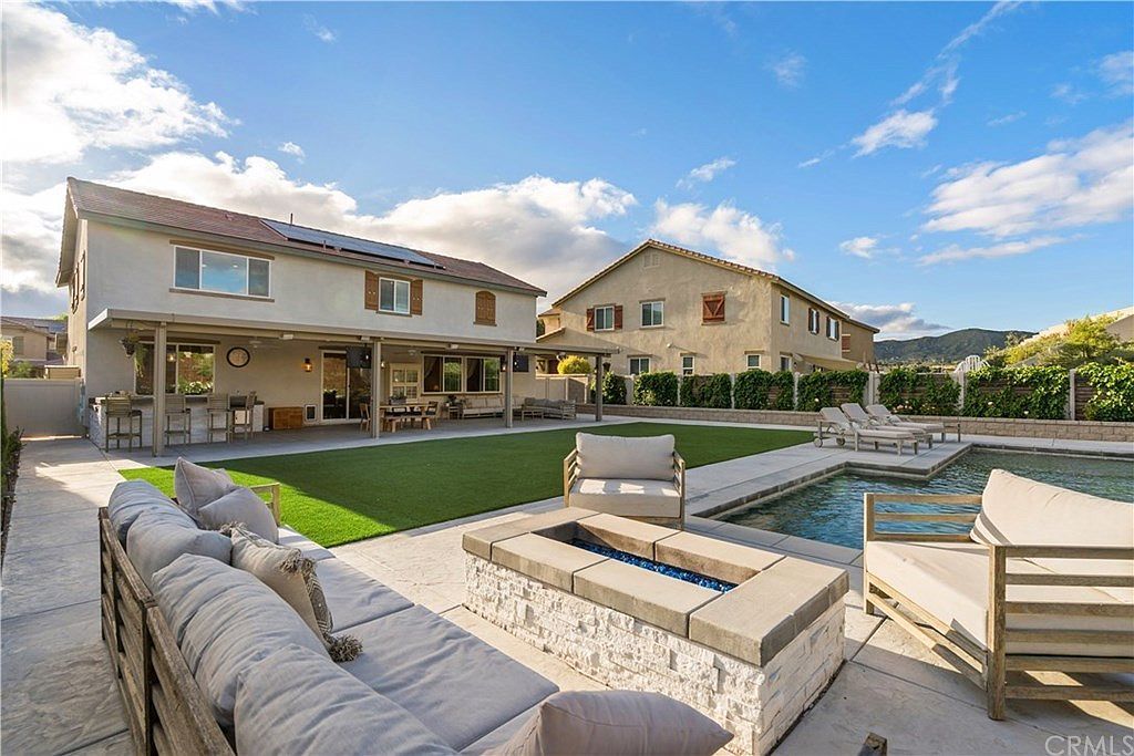 Backyard patio with a stone fire pit, outdoor furniture, a swimming pool, and two multi-story houses under a blue sky.