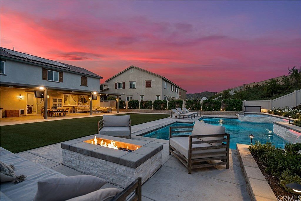 A backyard patio with a lit fire pit and outdoor seating next to a swimming pool at sunset with a pink and orange sky.
