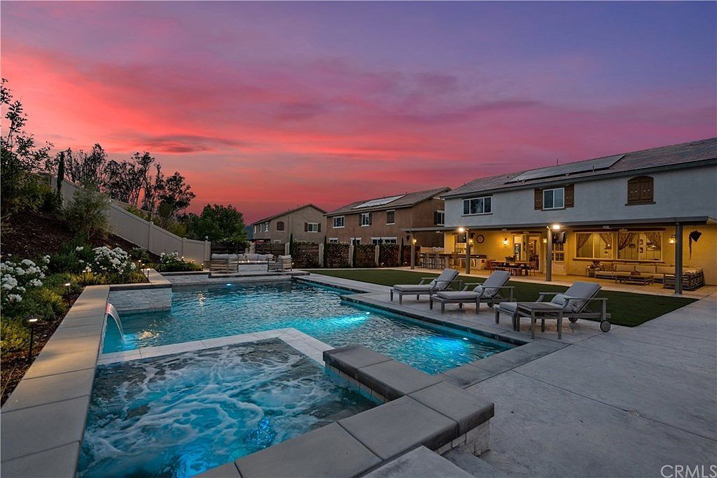 A backyard swimming pool and hot tub at twilight with glowing blue lights, lounge chairs, and a house in the background.