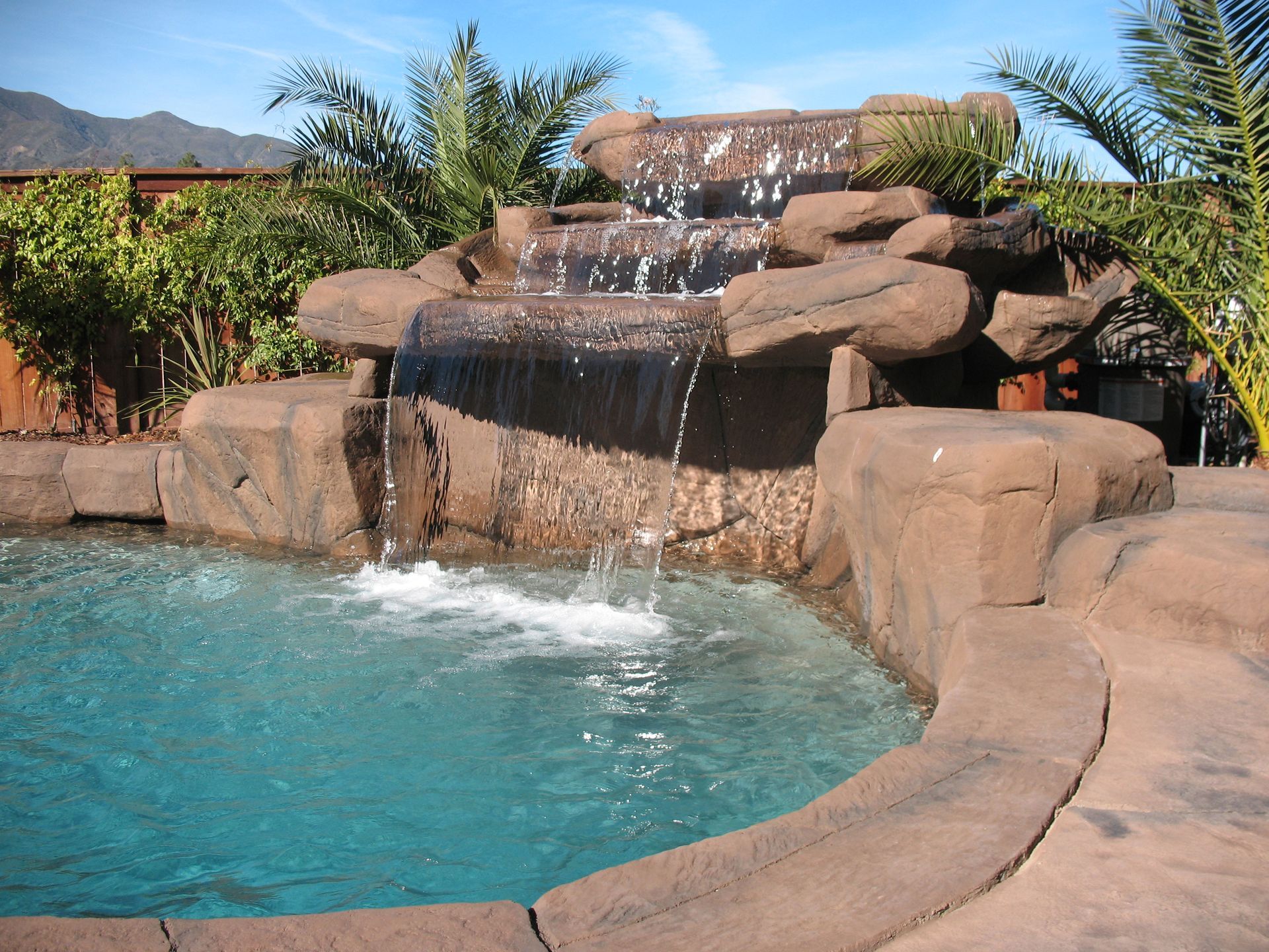 A multi-tiered artificial rock waterfall cascades into a clear blue swimming pool surrounded by greenery.