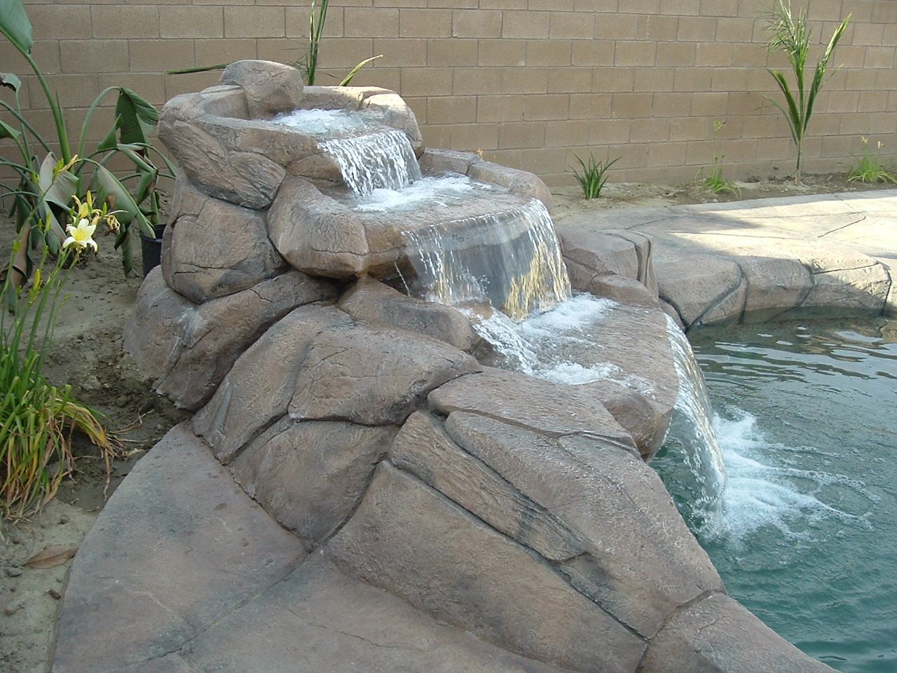 A tiered stone waterfall cascades into a swimming pool beside a brick wall and green foliage.