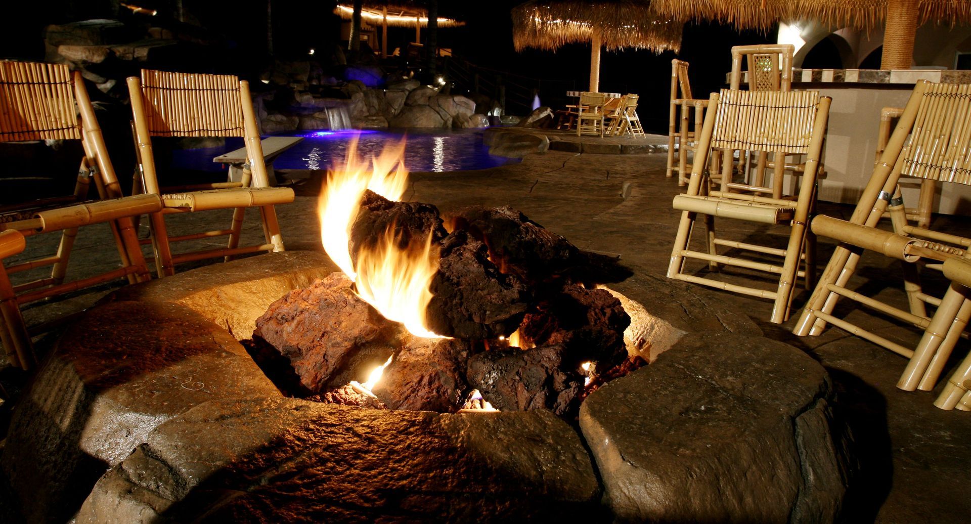 A fire pit with glowing flames set in a stone patio, surrounded by bamboo-style chairs under an outdoor tiki-style bar.