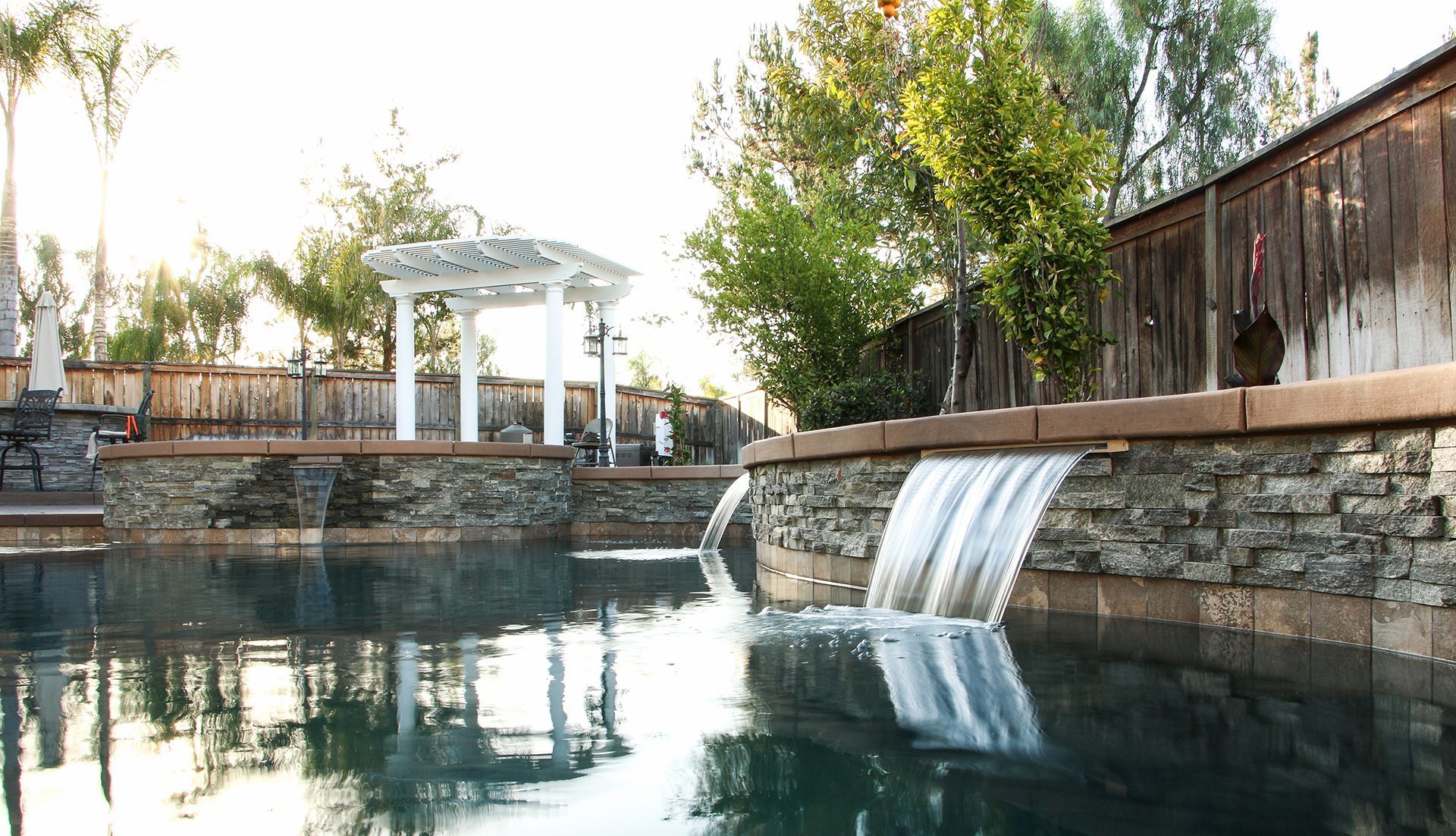 A backyard pool features stone retaining walls with cascading waterfalls, a white pergola, and lush green trees.