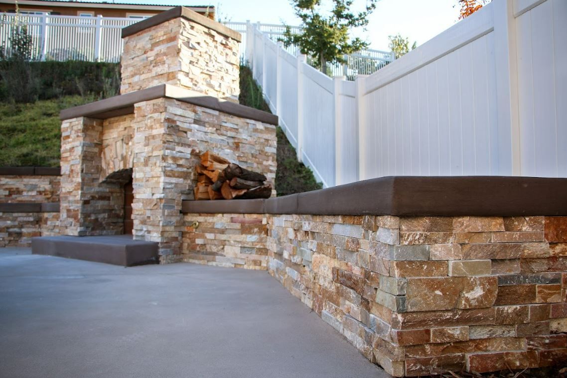 An outdoor stone fireplace and matching retaining wall on a concrete patio in front of a white fence.