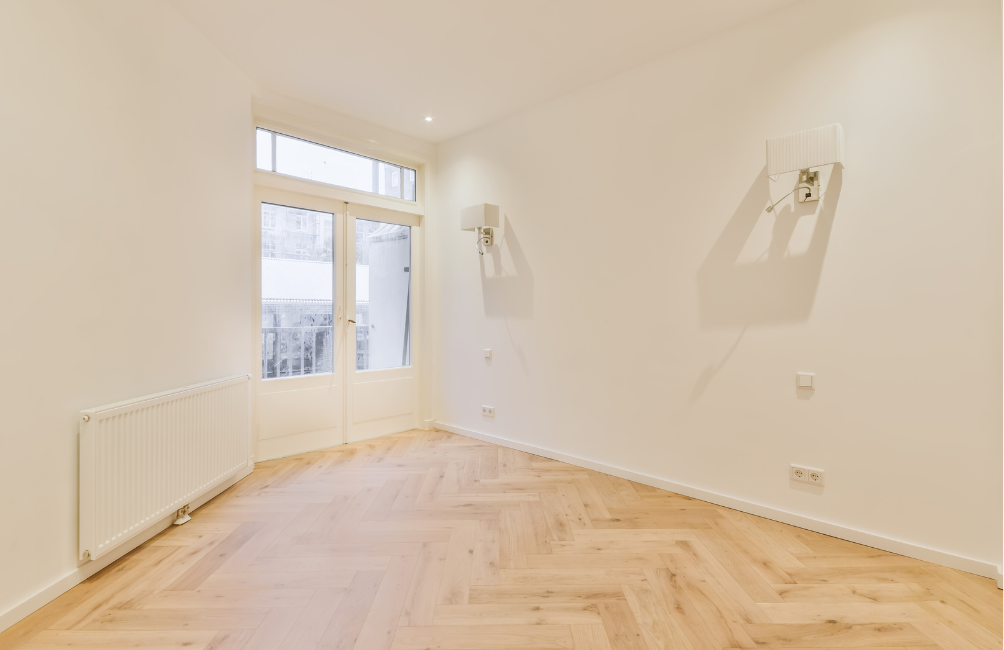 Pièce vide avec parquet clair, murs blancs et porte donnant sur un balcon. Un radiateur est fixé au mur de gauche.