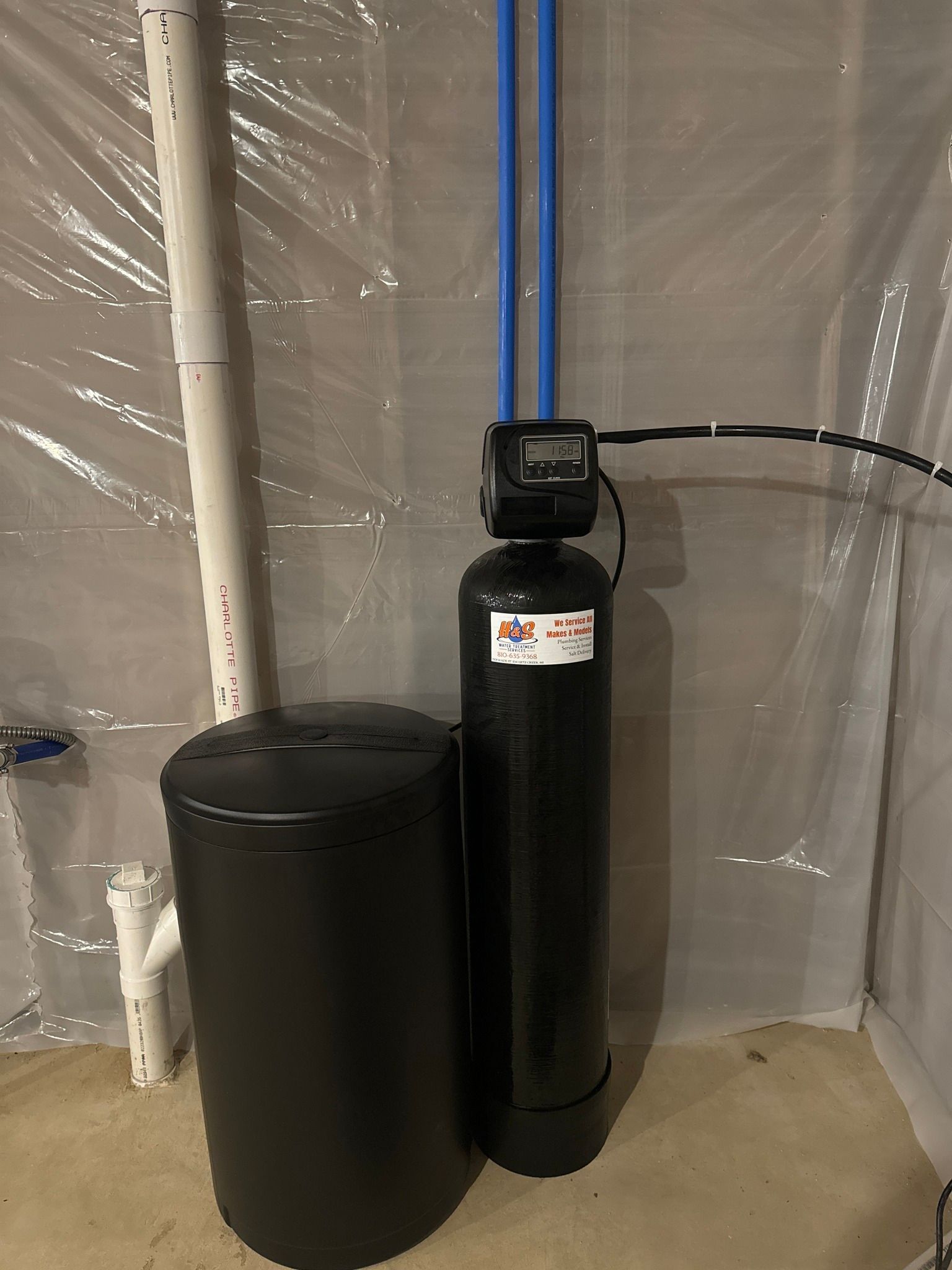 A black water softener is sitting next to a black tank in a basement.