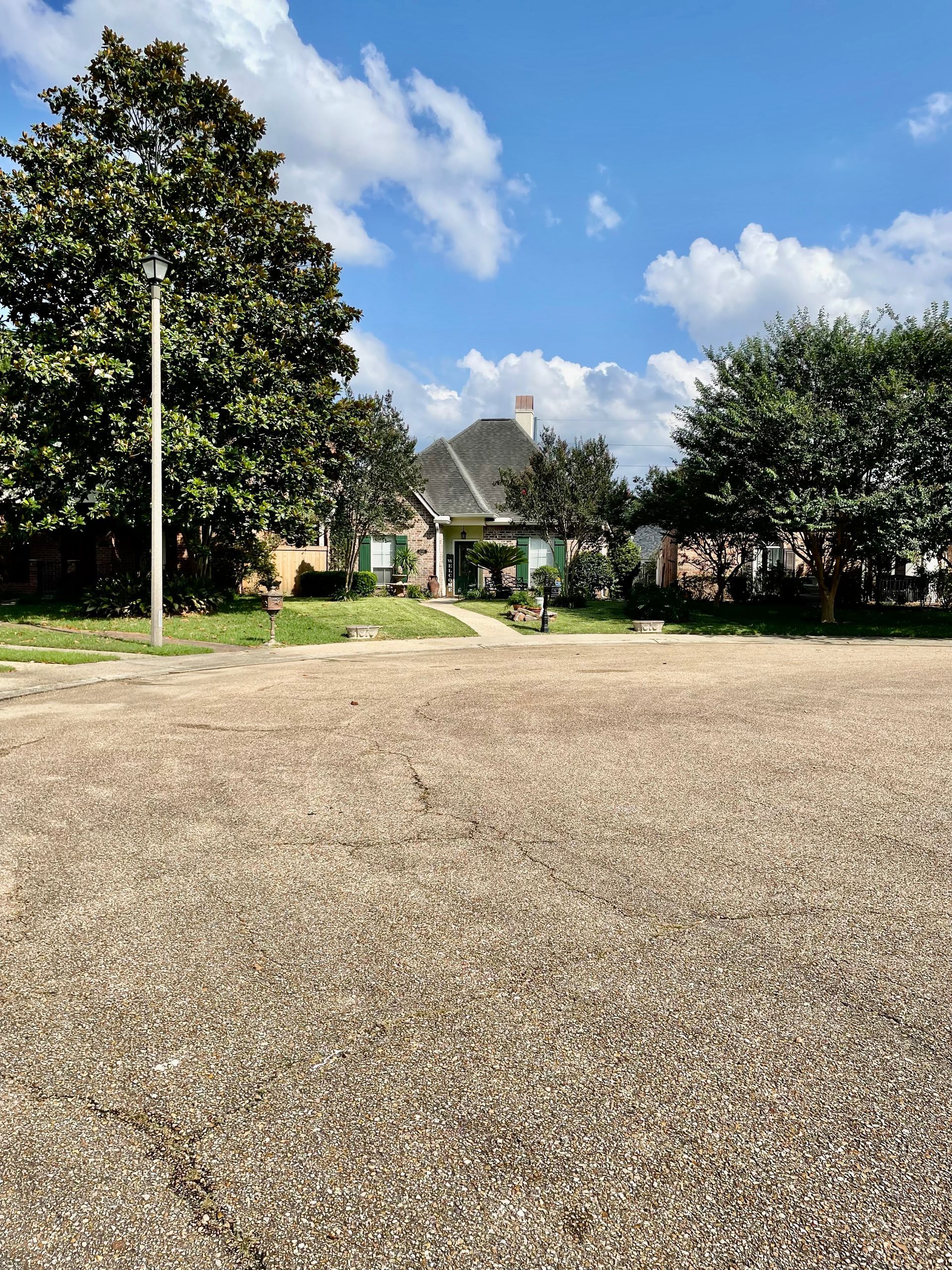 House with green shutters and a brown roof, trees on either side under a blue sky, fronted by a gravel area.
